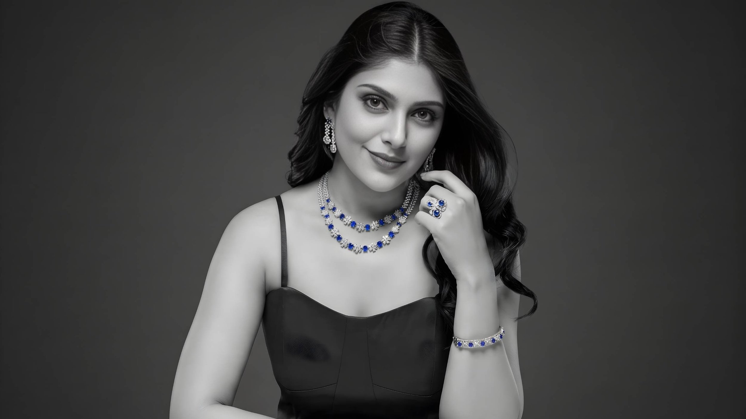 A woman wearing a black dress and jewelry, including earrings, necklaces, a ring, and a bracelet with blue and white stones, posing against a plain background.