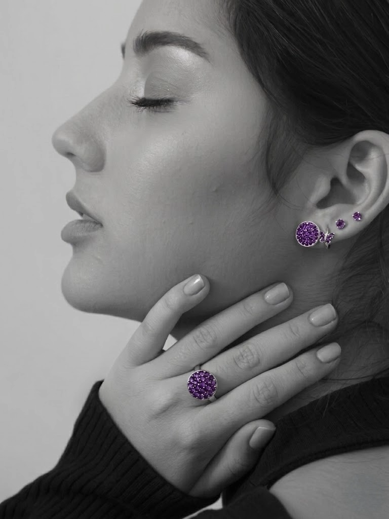 A woman wearing purple gemstone jewelry, including earrings and a ring, is shown in a black-and-white close-up portrait with a focus on her face, hand, and jewelry.