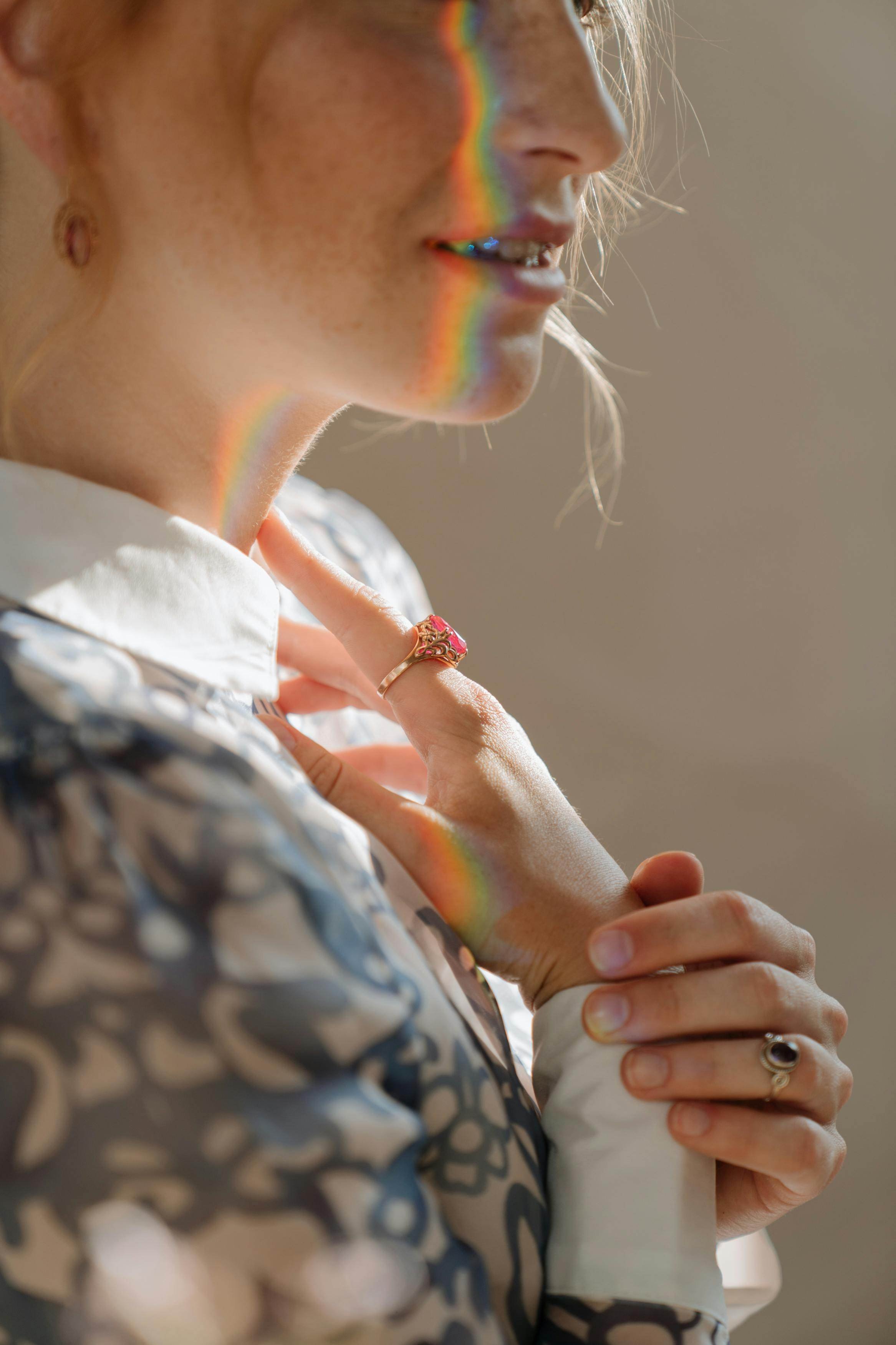 Close-up of a woman with rainbow light reflections on her face and hand, wearing rings, holding her arm and casually looking down.