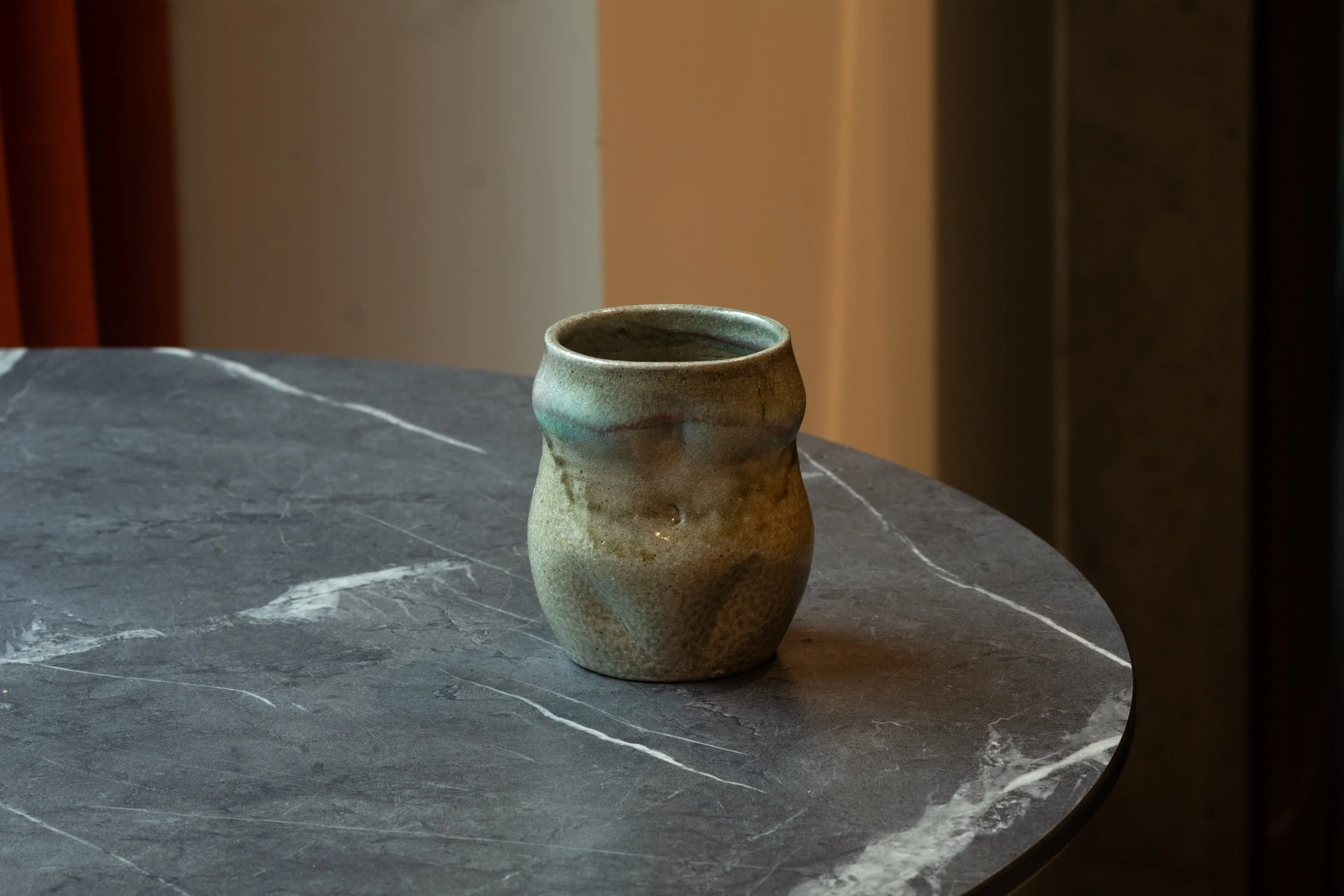 A small, rustic ceramic mug with a textured surface sits on a dark gray marble table with white veining