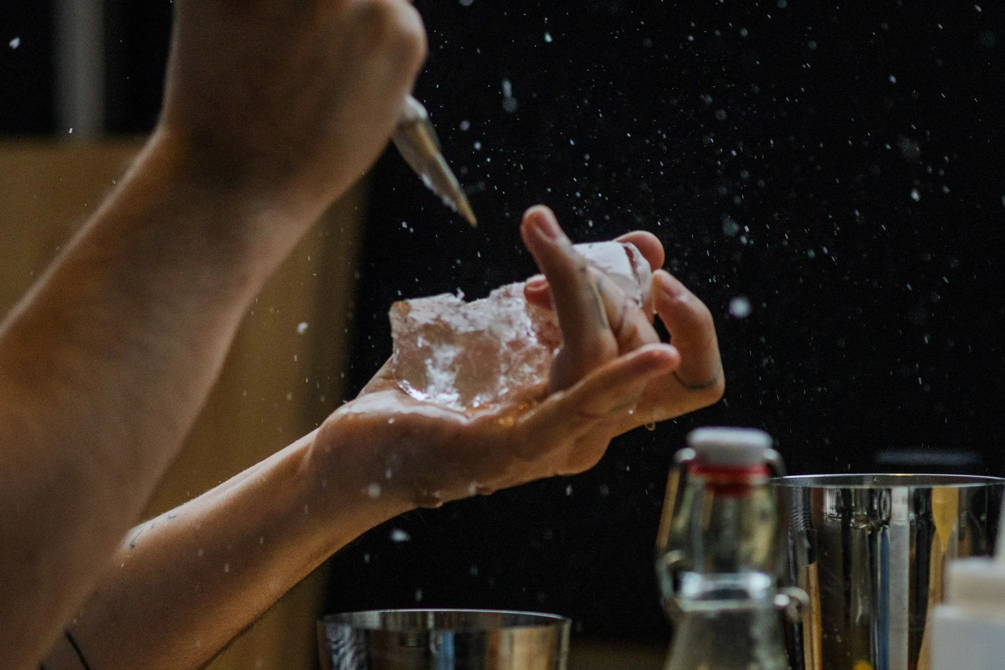 A person is holding a piece of ice in one hand while carving or shaping it with a tool in the other hand, with water splashing around in a dark background.
