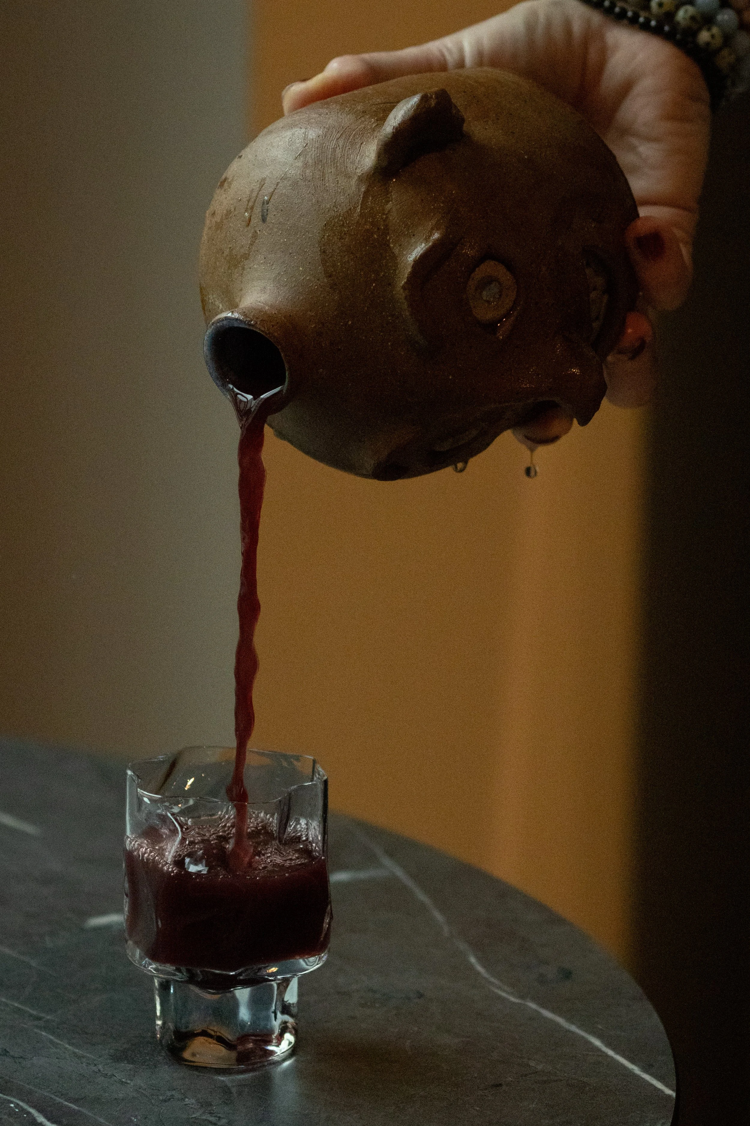 A person pouring red juice or wine from a brown ceramic vessel into a small glass.