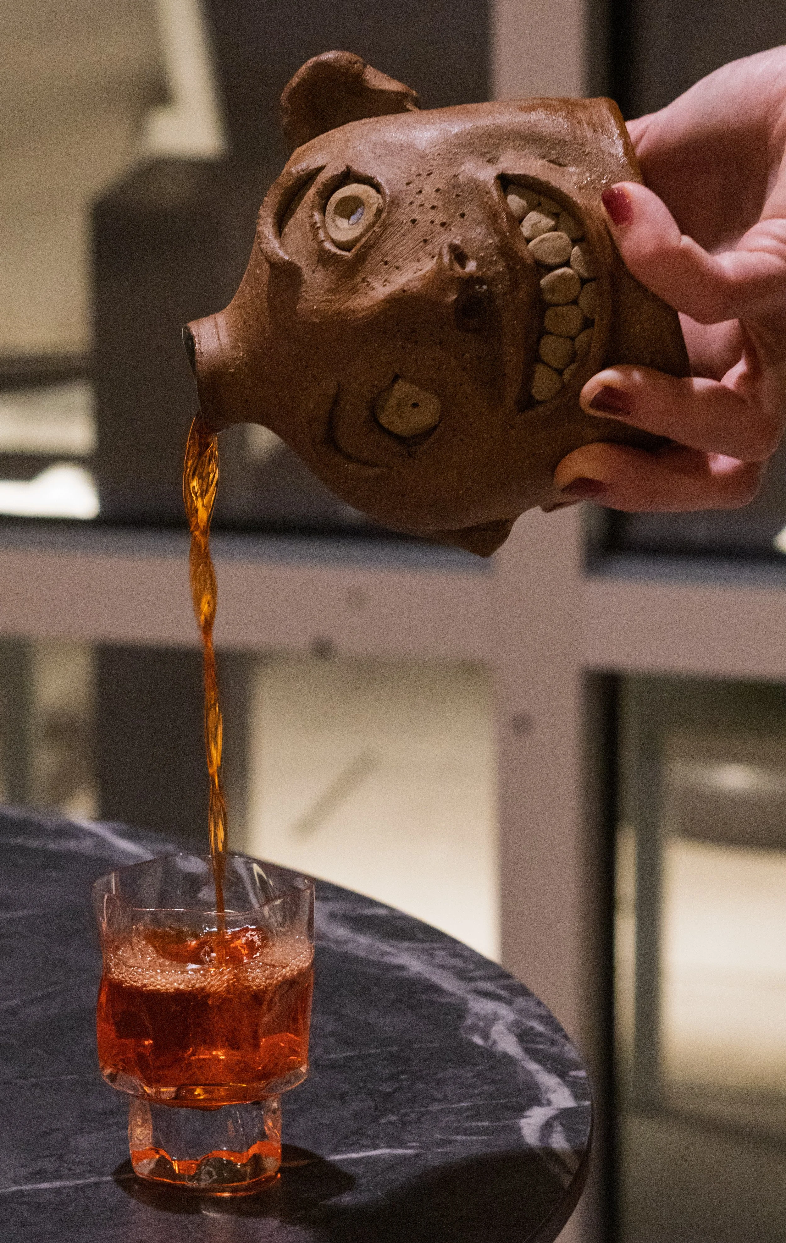 A person pouring a drink from a bear-shaped teapot into a glass. The teapot has a face with a large open mouth, one eye, and a snout, and appears to be made of ceramic.