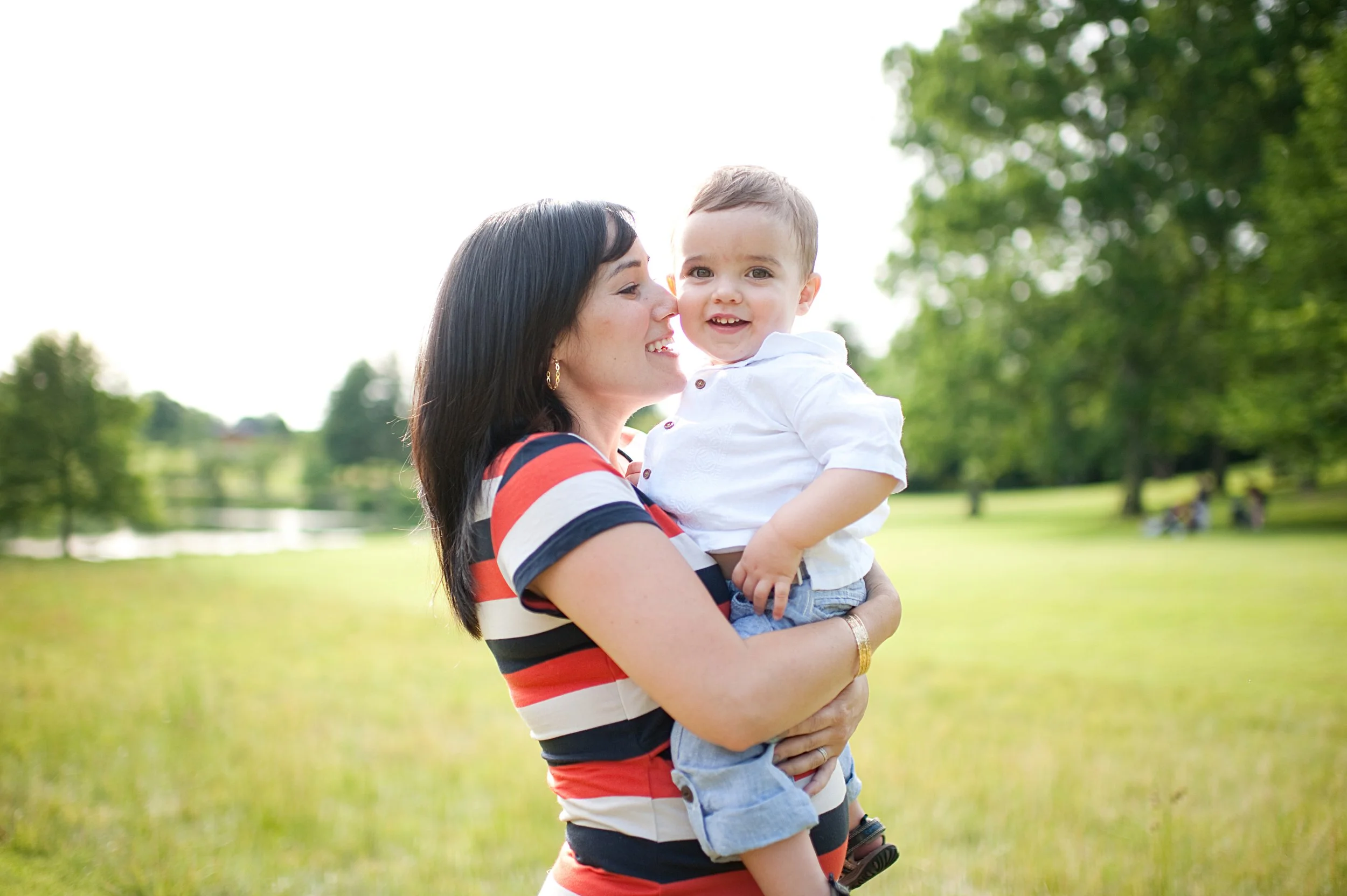 A woman holding a young boy outdoors in a park with green grass and trees in the background