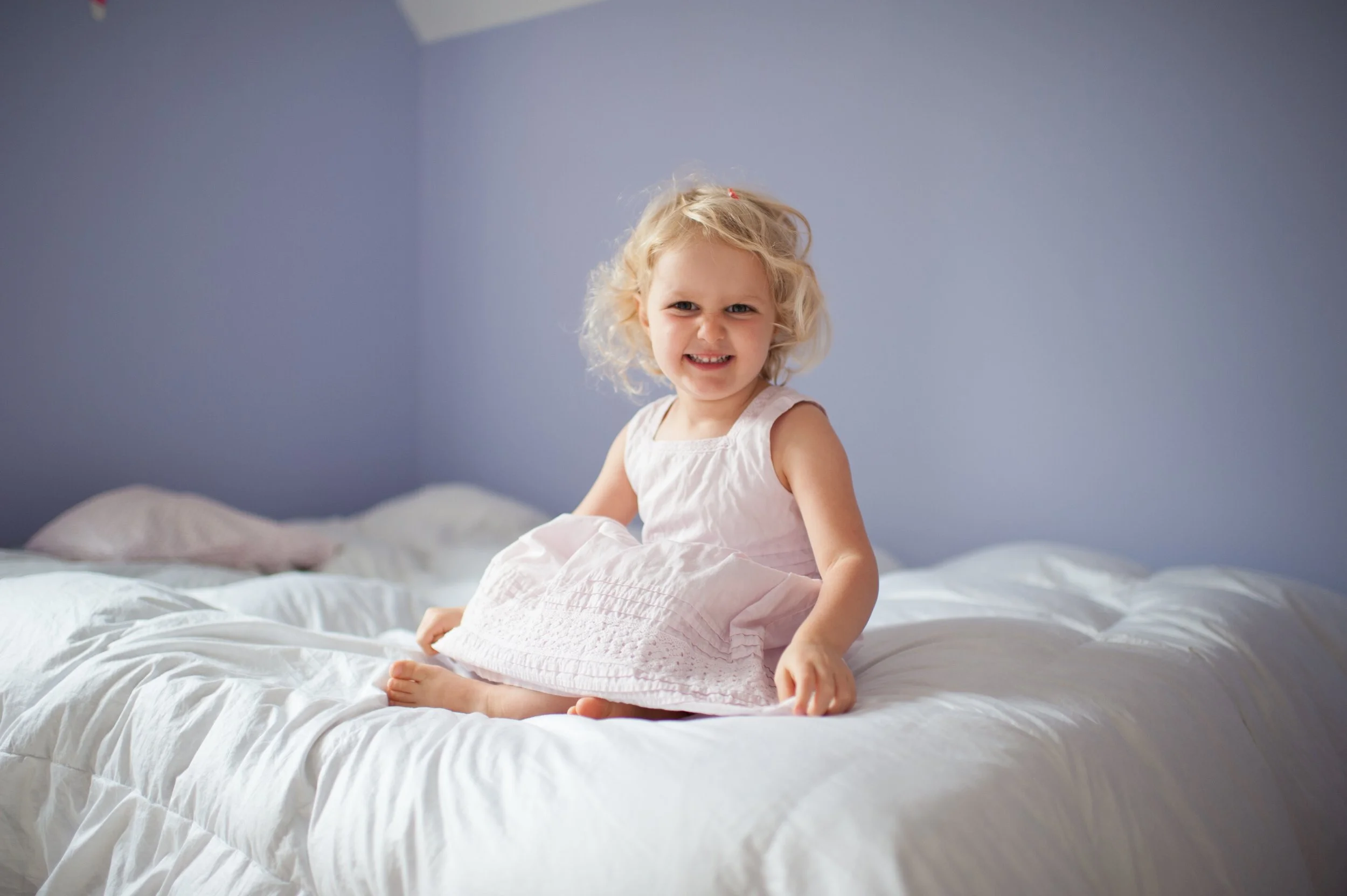 A young girl with blonde curly hair sitting on a white bed, smiling at the camera.
