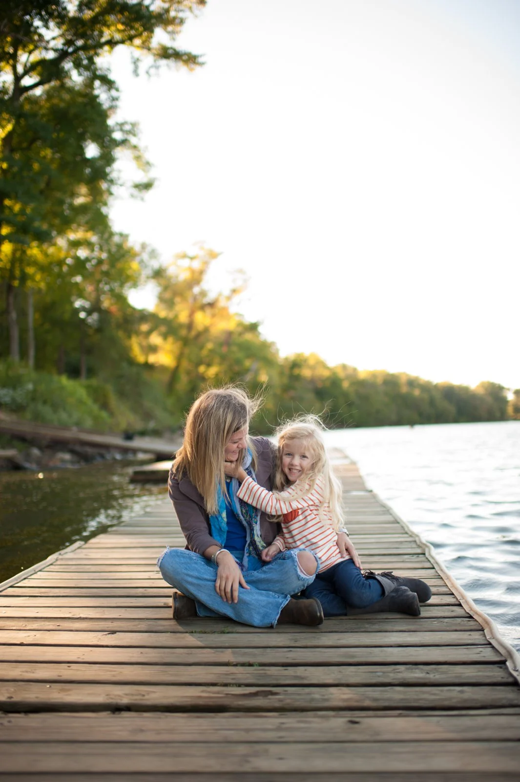 A woman and a young girl sitting on a wooden dock by a lake, laughing and playing together with trees in the background during sunset.