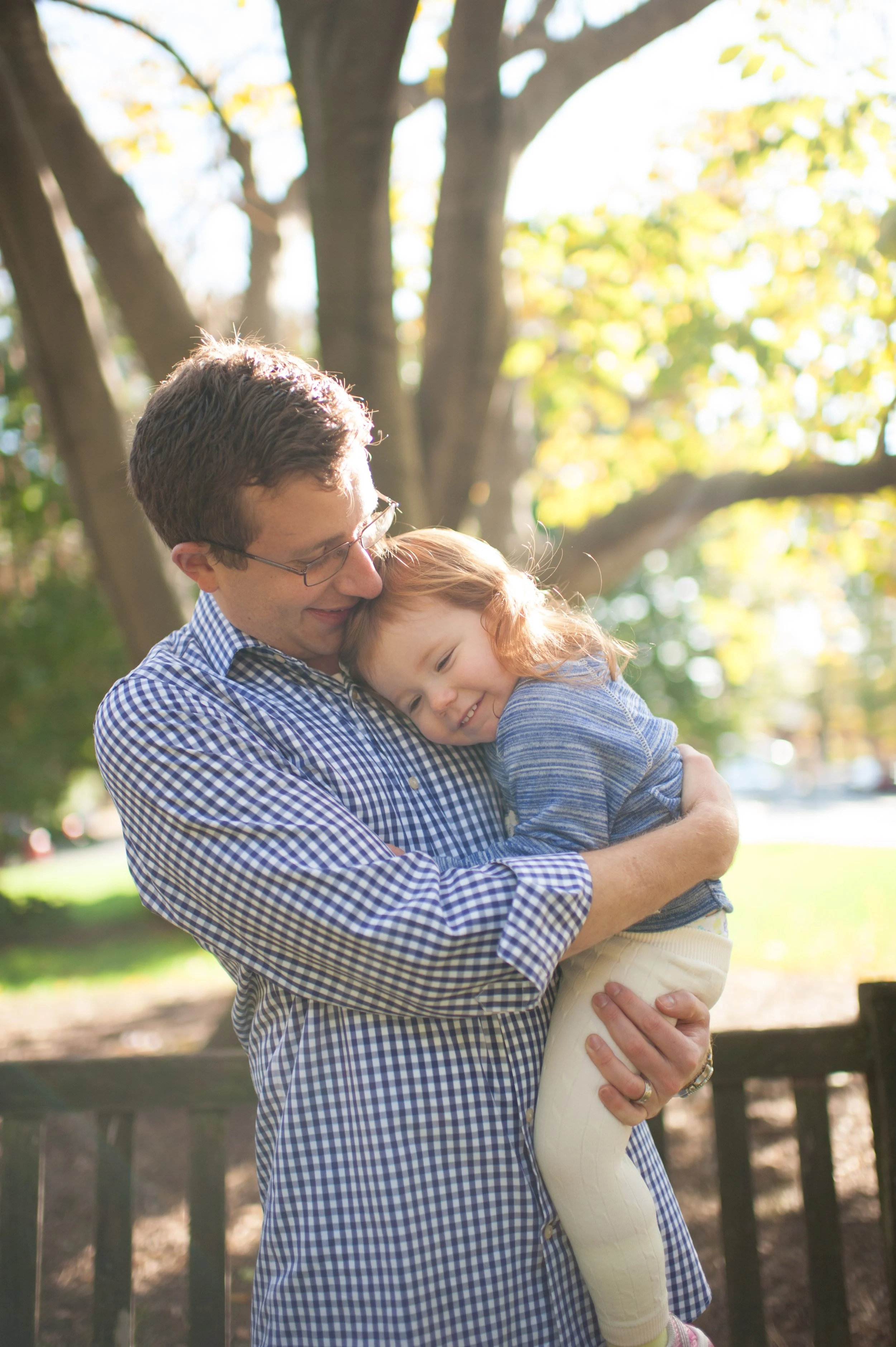 A man holding a young girl in an outdoor park with mature trees in the background, both smiling as they enjoy the moment.