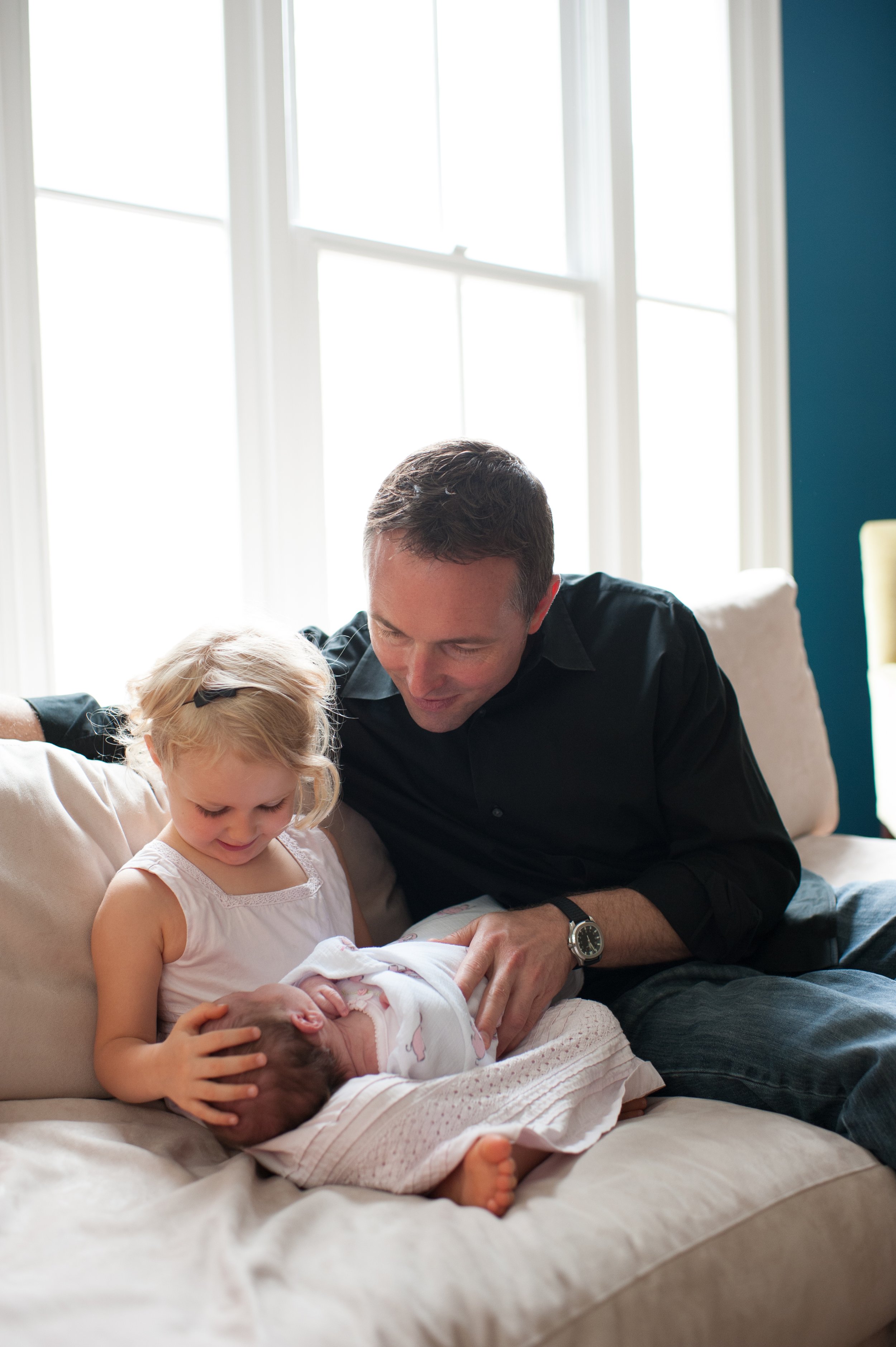 A man, a young girl, and a newborn baby girl sitting on a beige couch in front of large windows with sunlight, the girl is looking at the baby.