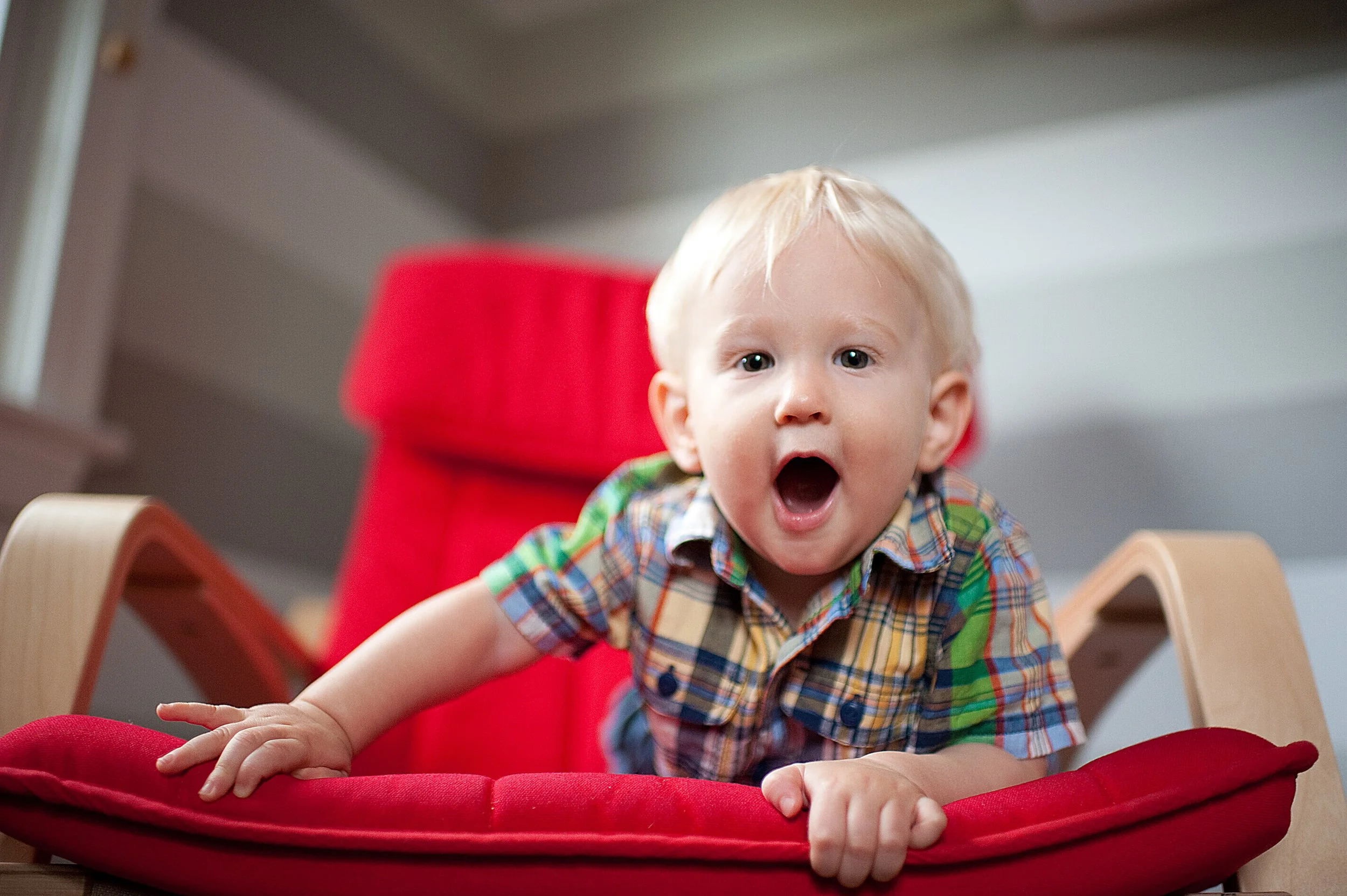 A young boy with blonde hair and wearing a colorful plaid shirt is leaning on a red cushion in a wooden chair, with a surprised or excited expression on his face.