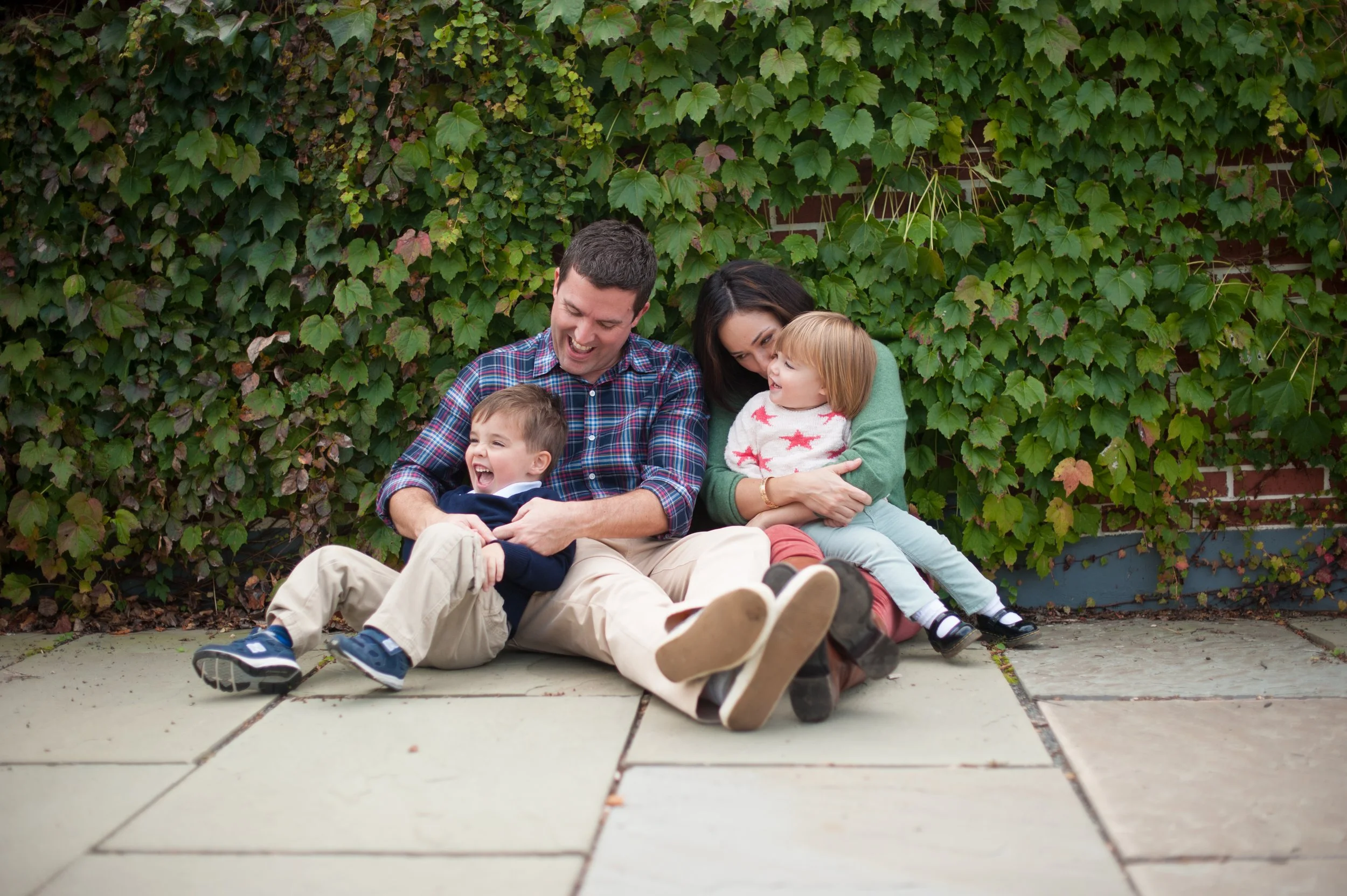 Family of four sitting on sidewalk in front of green leafy wall, playing and laughing together.