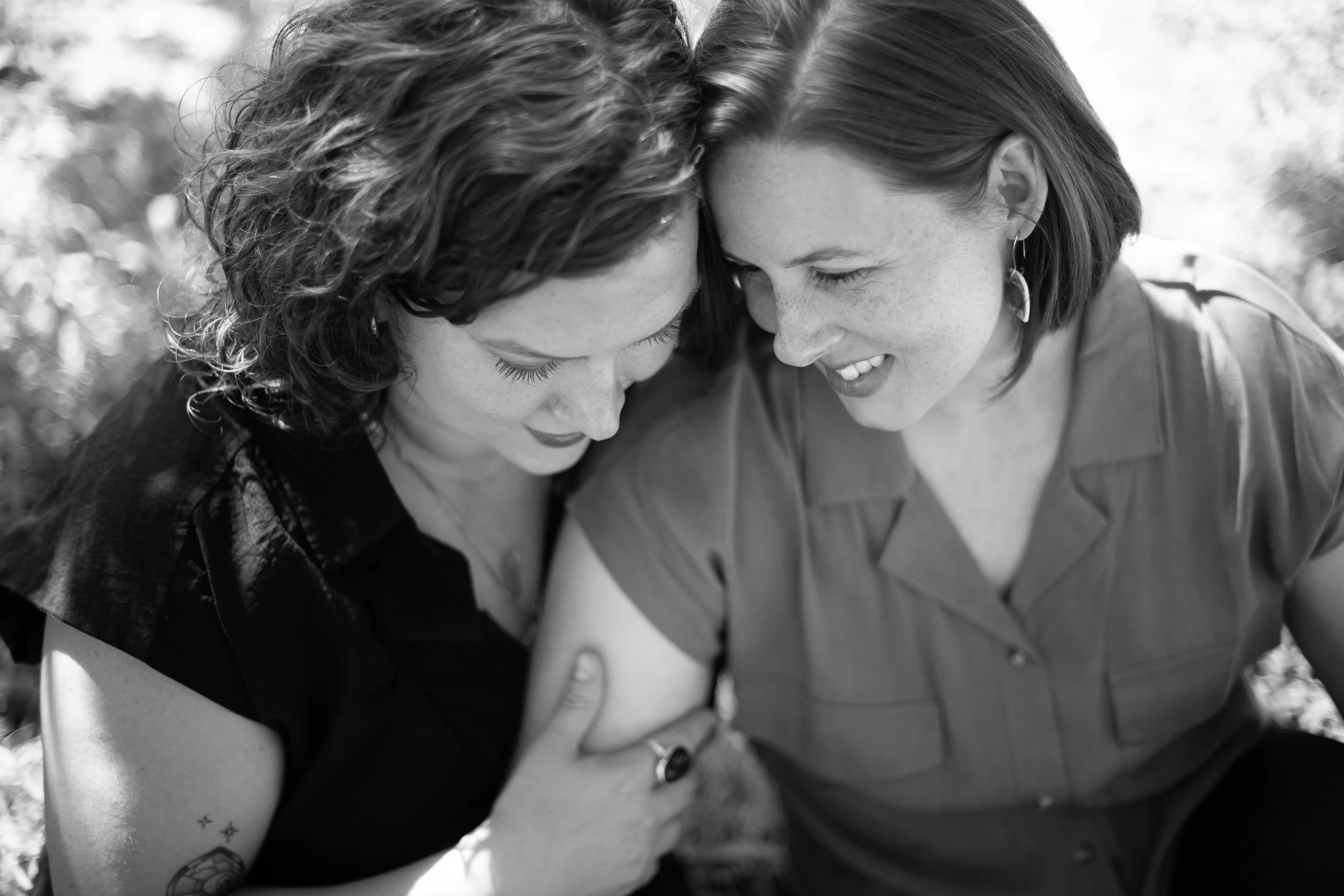 Two women with curly and straight hair closely facing each other with their foreheads touching, smiling warmly, outdoors in a black-and-white photo.