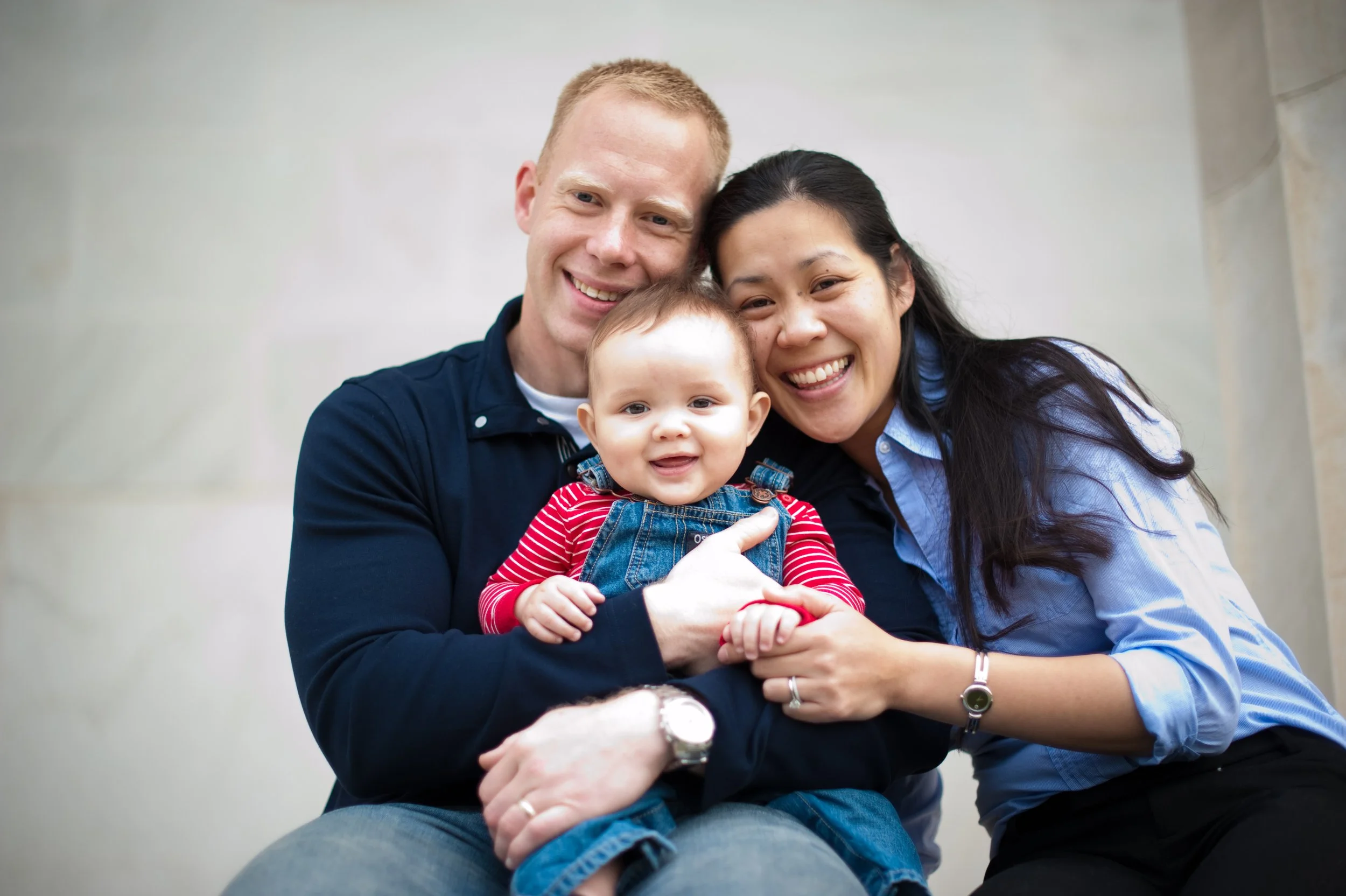 A happy family of three, including a man, woman, and a baby, smiling and sitting close together outside against a light-colored wall.
