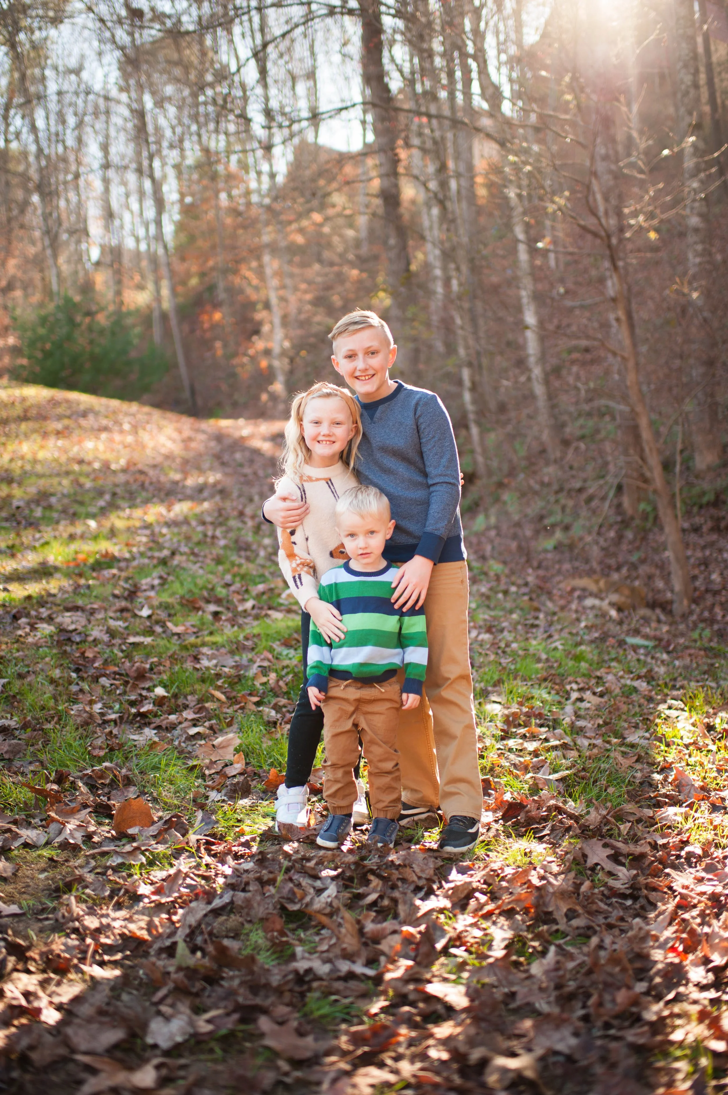 Three children standing outdoors in a wooded area during fall, with sunlight filtering through trees, surrounded by fallen leaves.