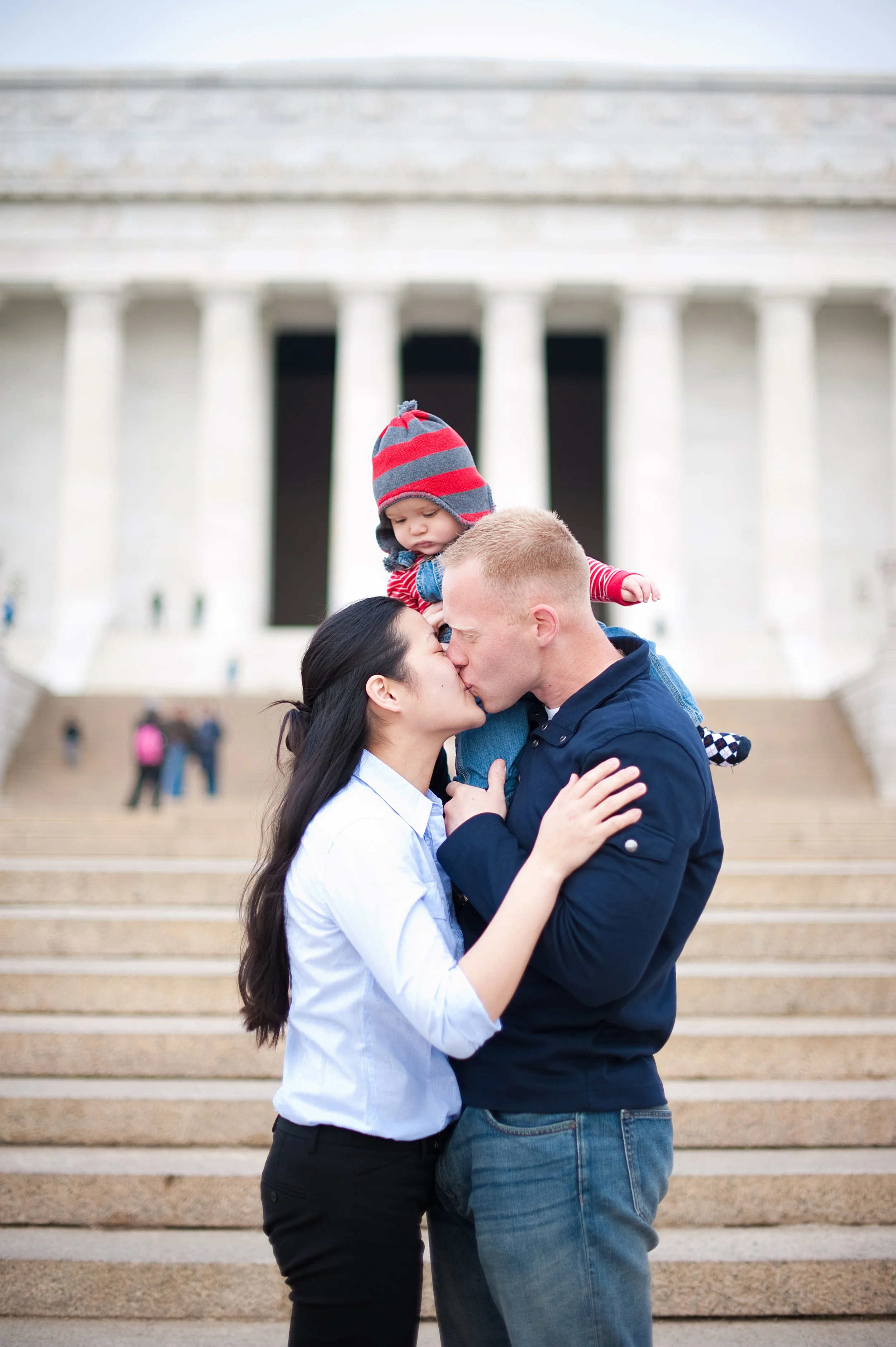 A couple kissing with a young child on his parents' shoulders, at the Lincoln Memorial in Washington, D.C., with stairs and visitors in the background.