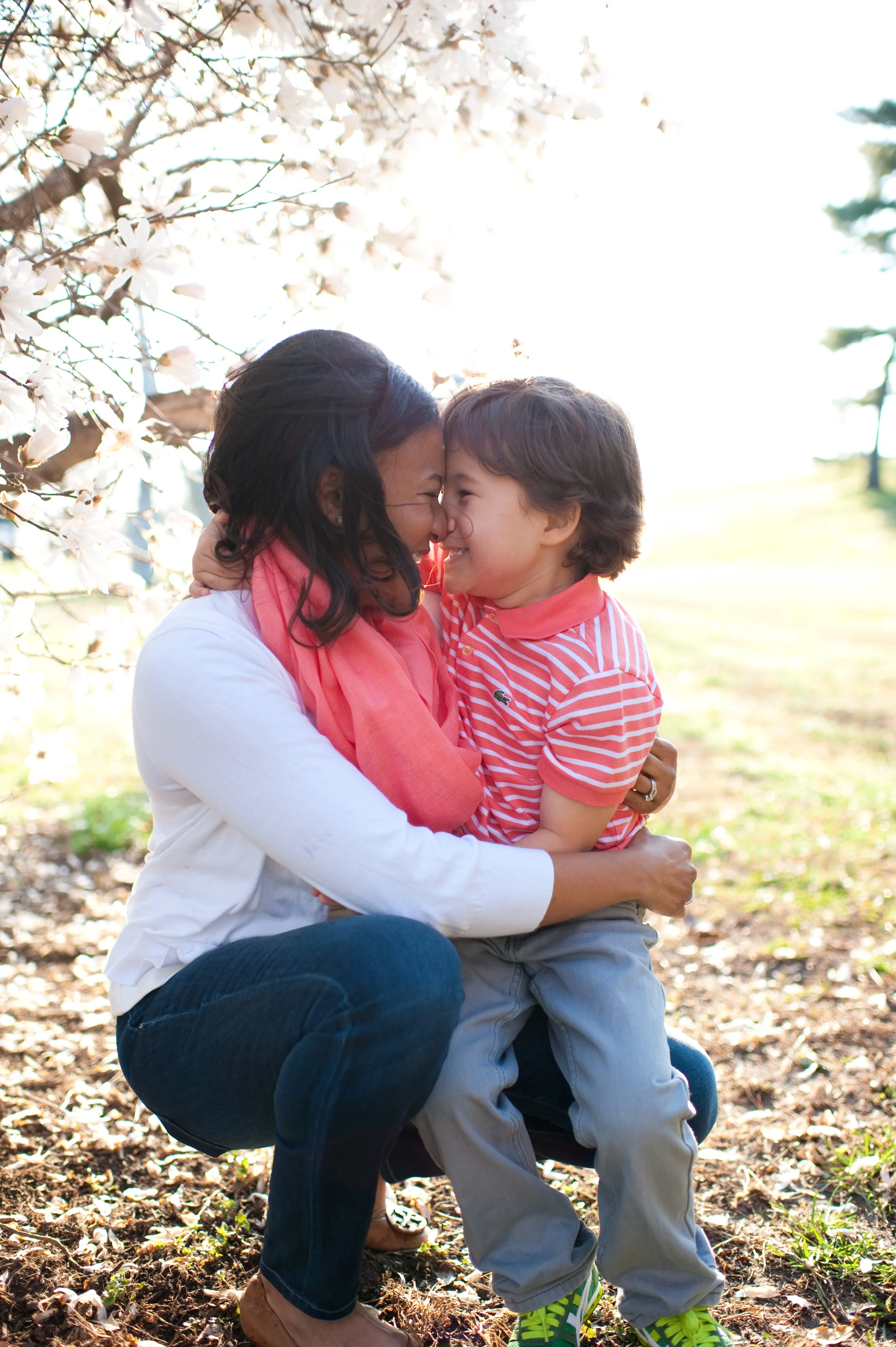 A woman and a young boy are smiling and touching foreheads, enjoying a joyful moment outdoors in a park with blooming trees.