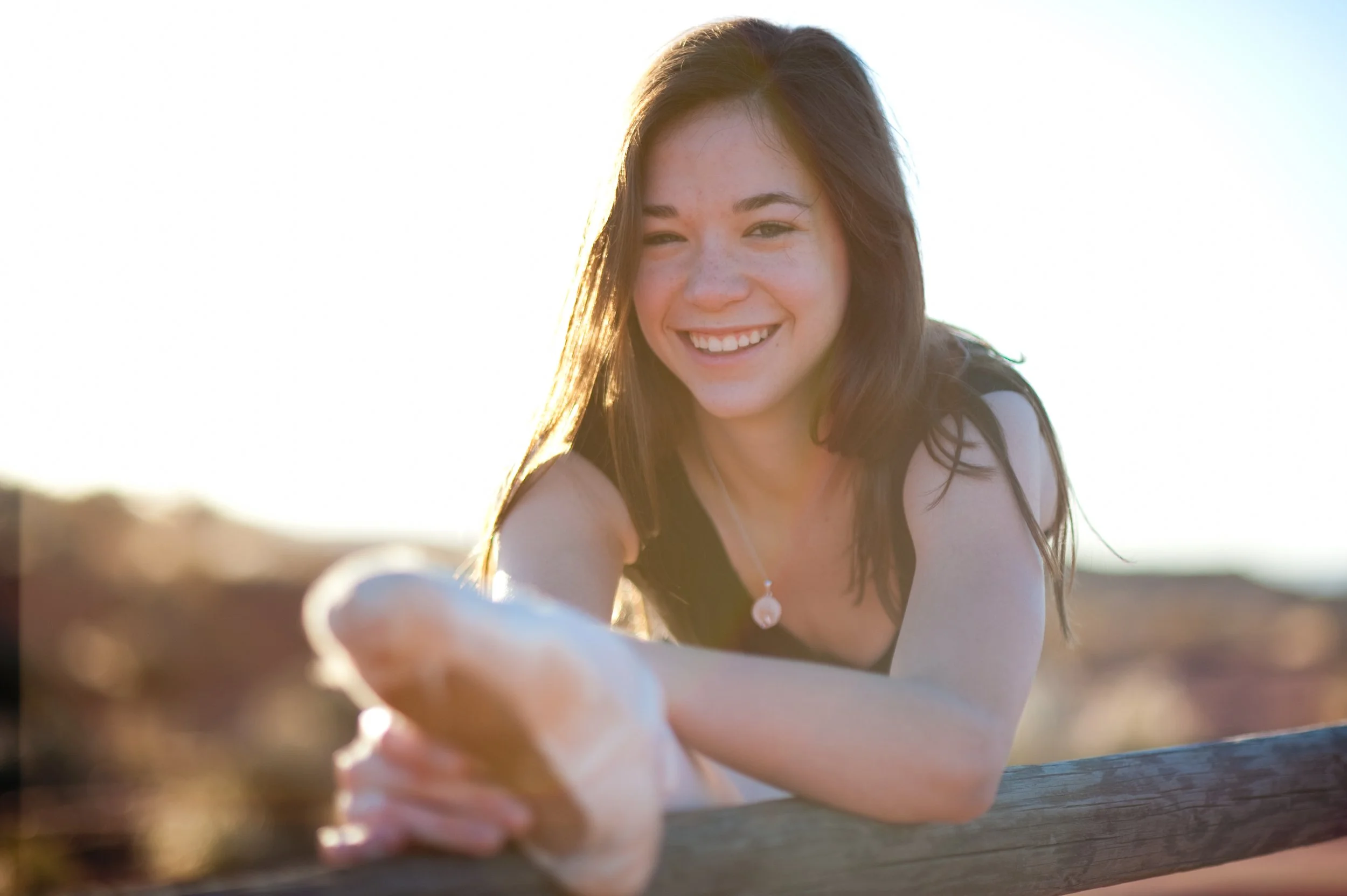 Young woman with dark hair smiling and reaching out toward the camera outdoors in bright sunlight.