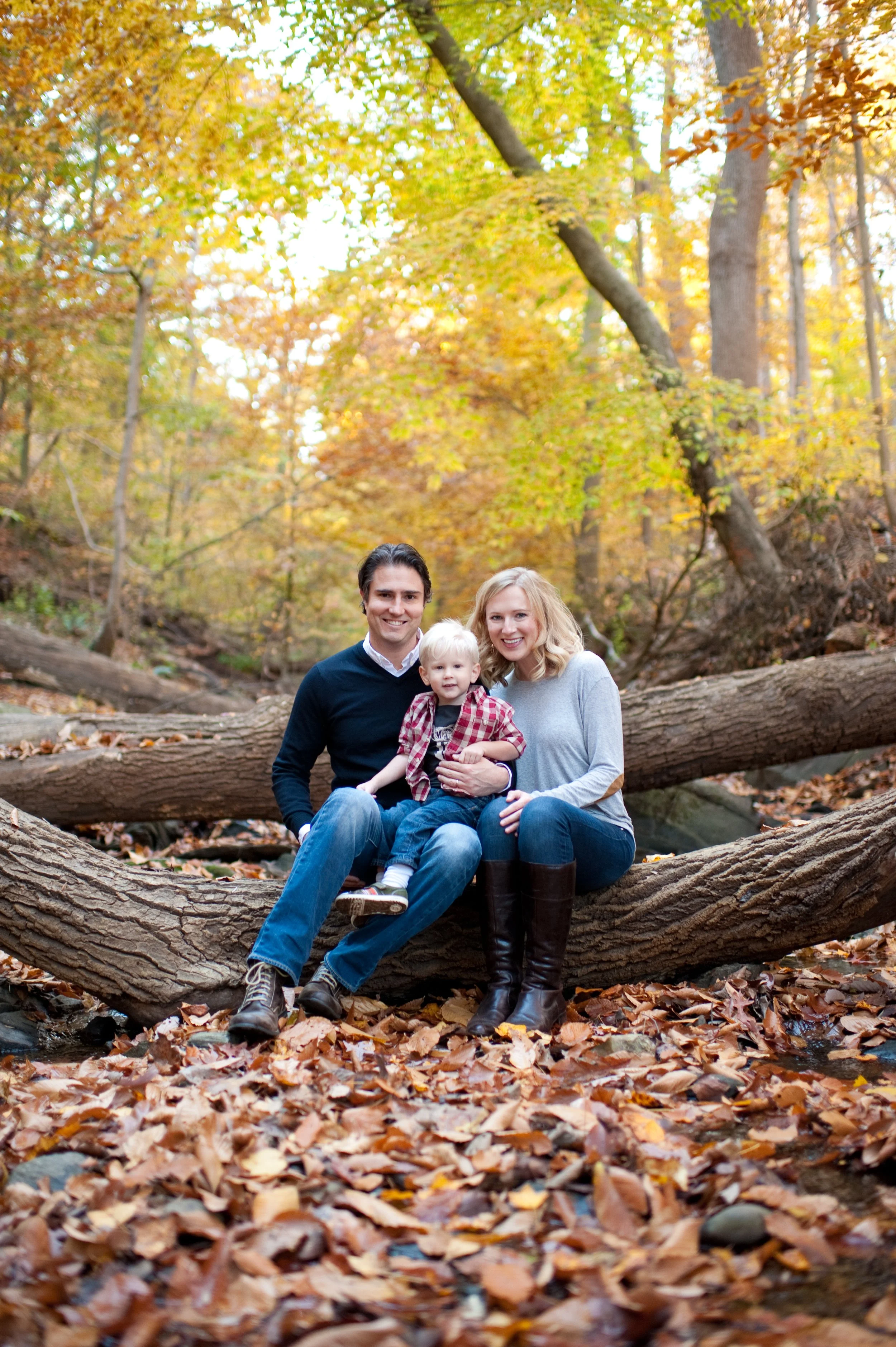 A family of three sitting on a fallen tree in an autumn forest, surrounded by colorful fall leaves. The father, mother, and young child are smiling at the camera.