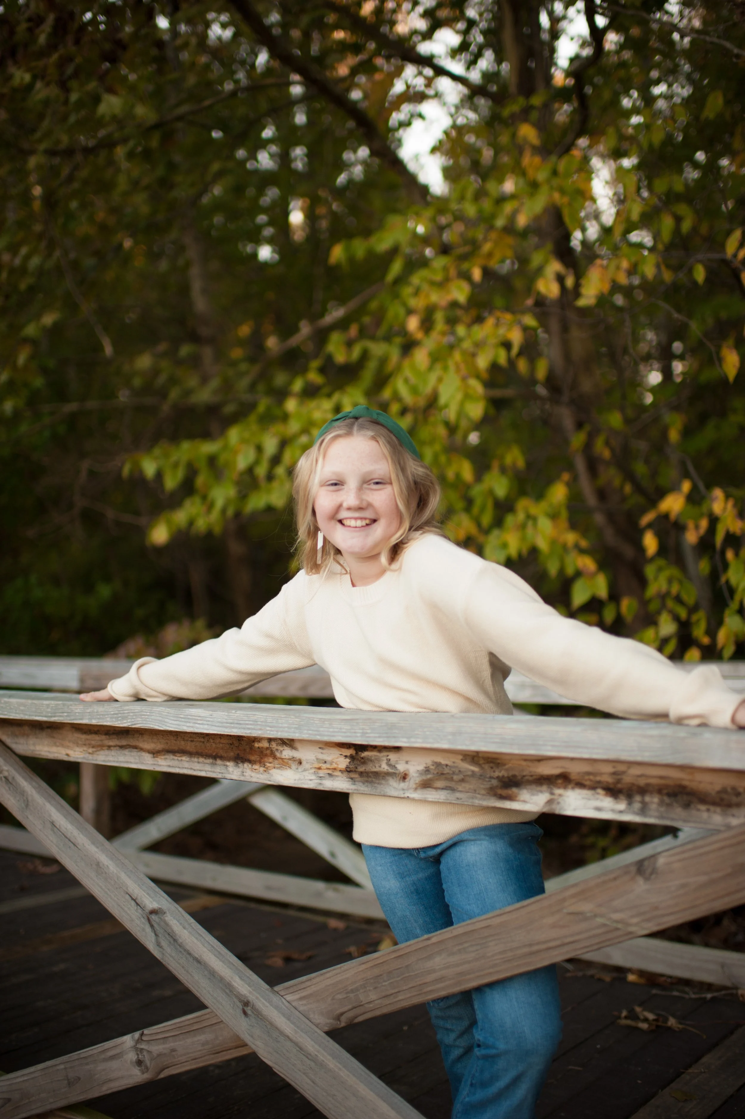A young girl with blonde hair smiling, wearing a cream sweater and blue jeans, standing outdoors on a wooden bridge surrounded by green and yellow autumn leaves.