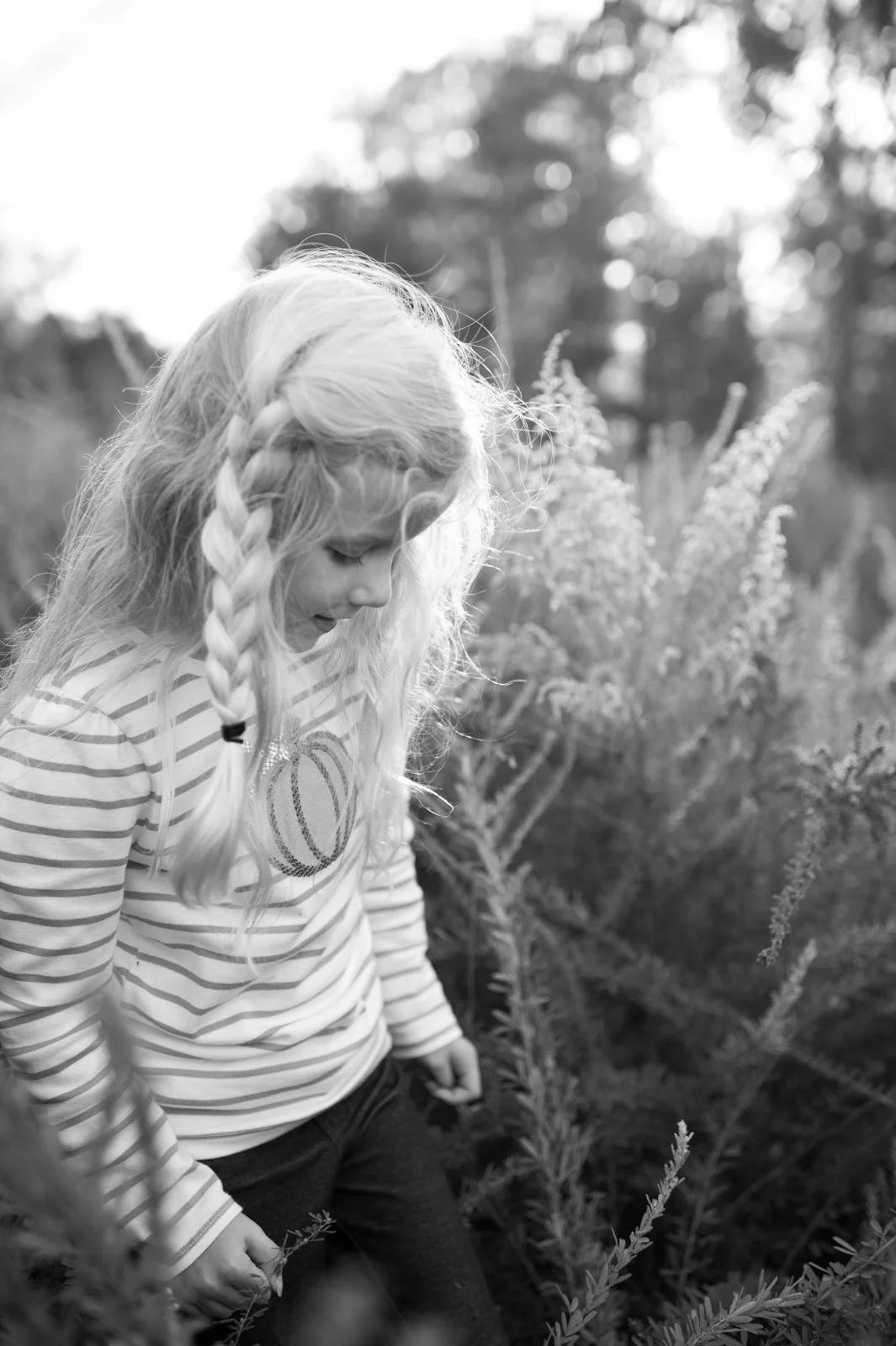 A young girl with long, braided blonde hair wearing a striped shirt outdoors among plants and trees in black and white.