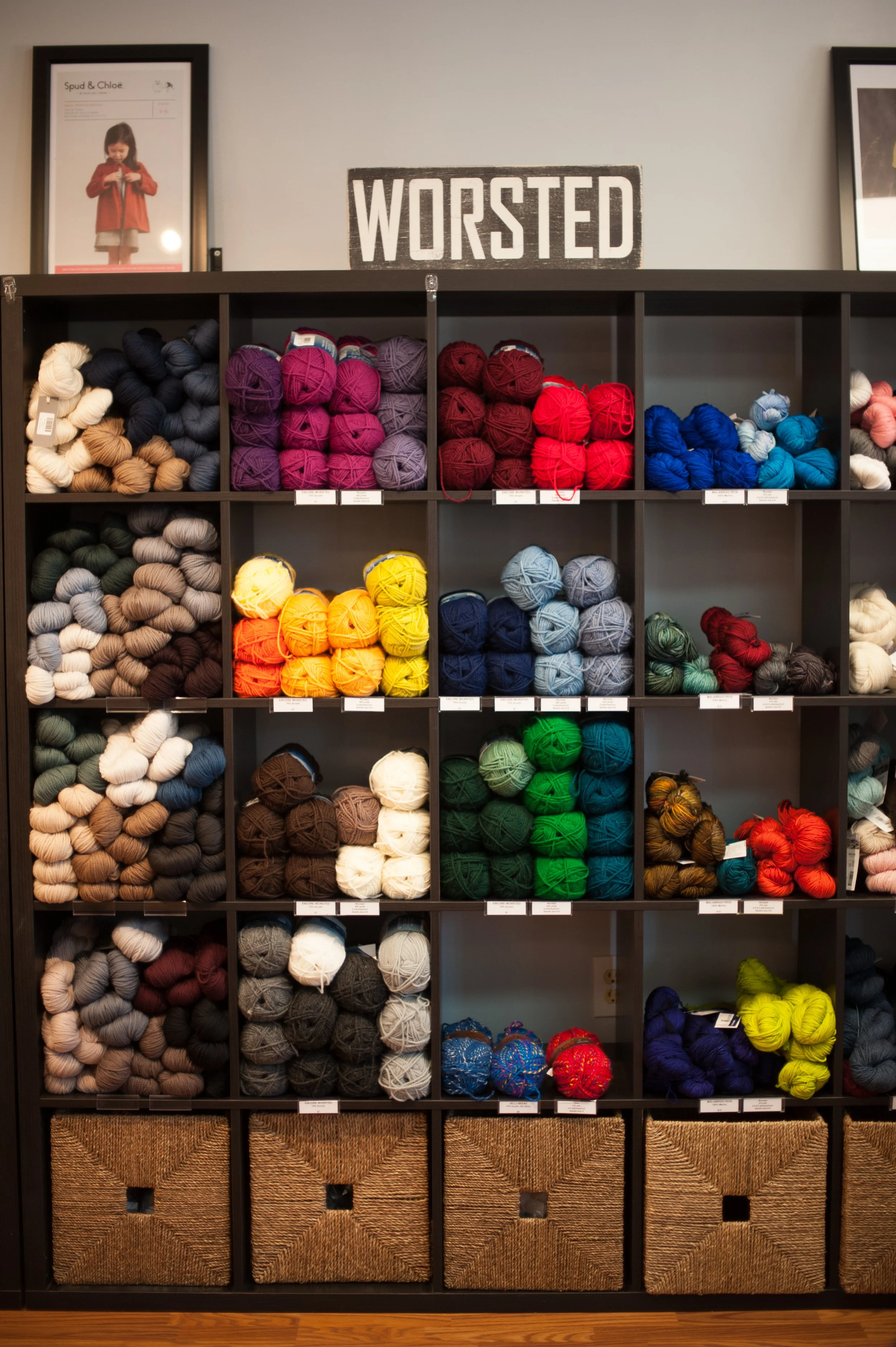 Colorful yarn balls arranged on a black cubical shelf with a sign reading 'WORSTED' on top, and framed pictures on the wall behind the shelf.