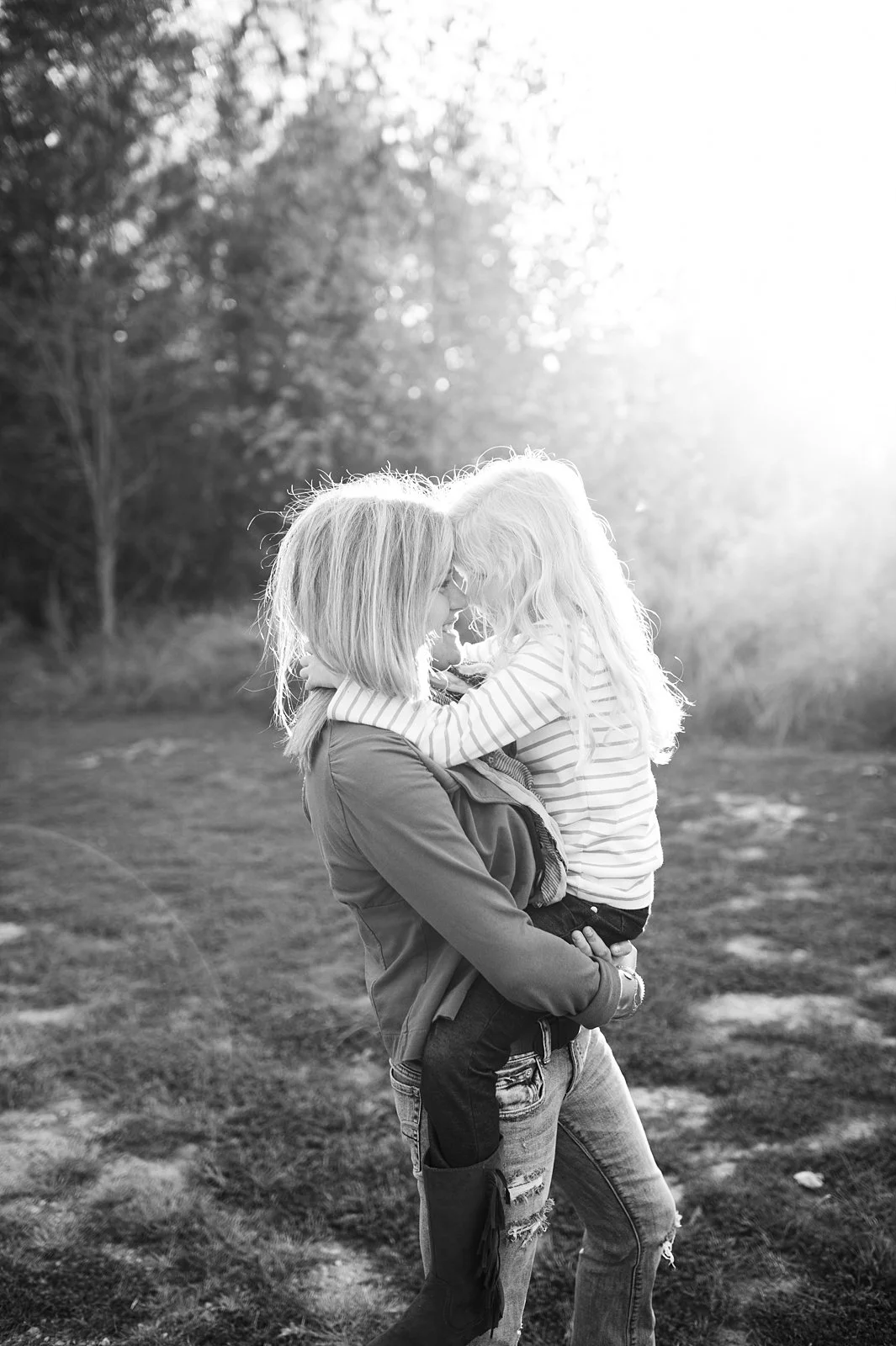 A woman is holding a young girl in an outdoor setting, both smiling and touching foreheads with sunlight in the background.