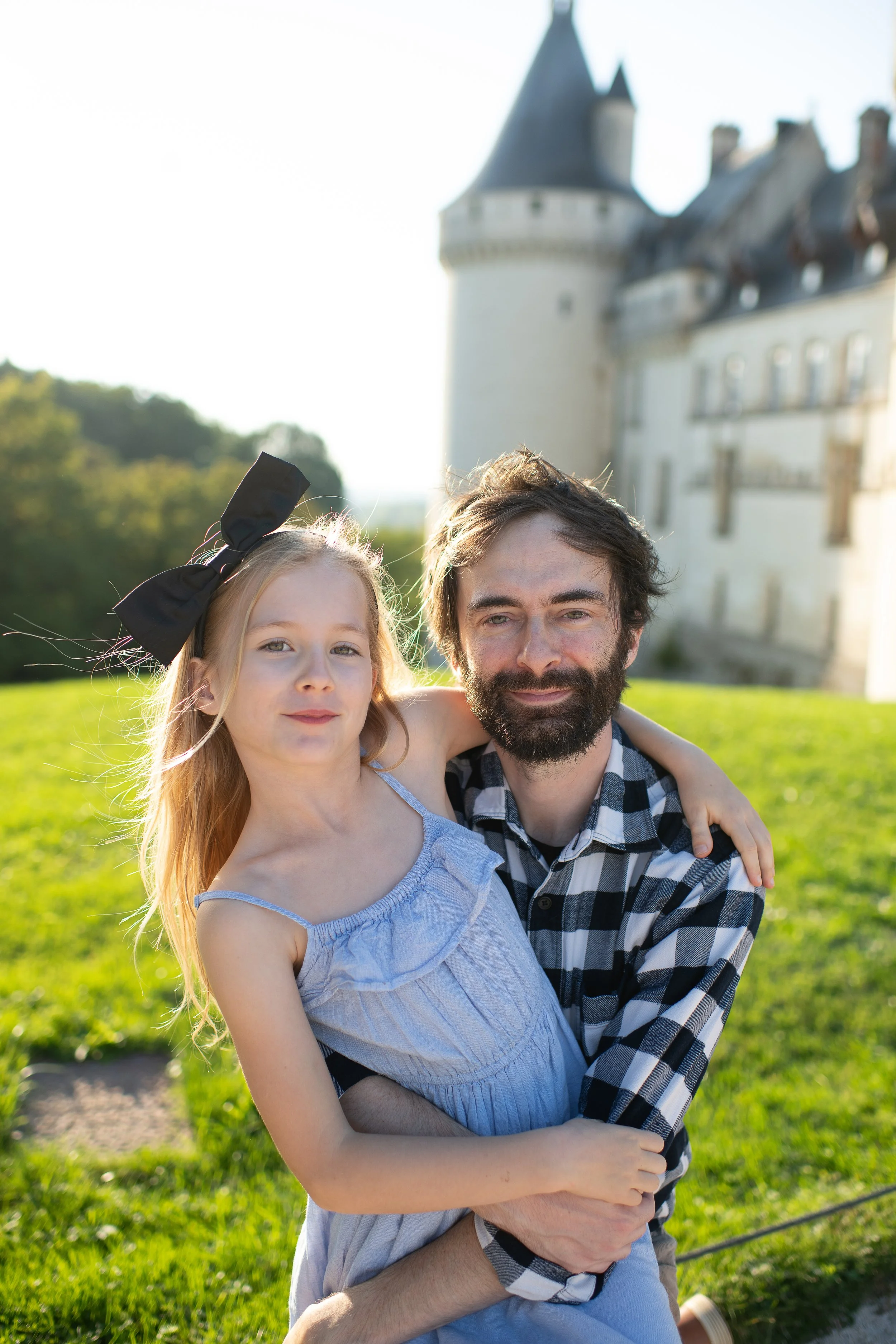 A man with a beard and a young girl with blonde hair and a large black bow in her hair, hugging each other outdoors with a castle in the background.