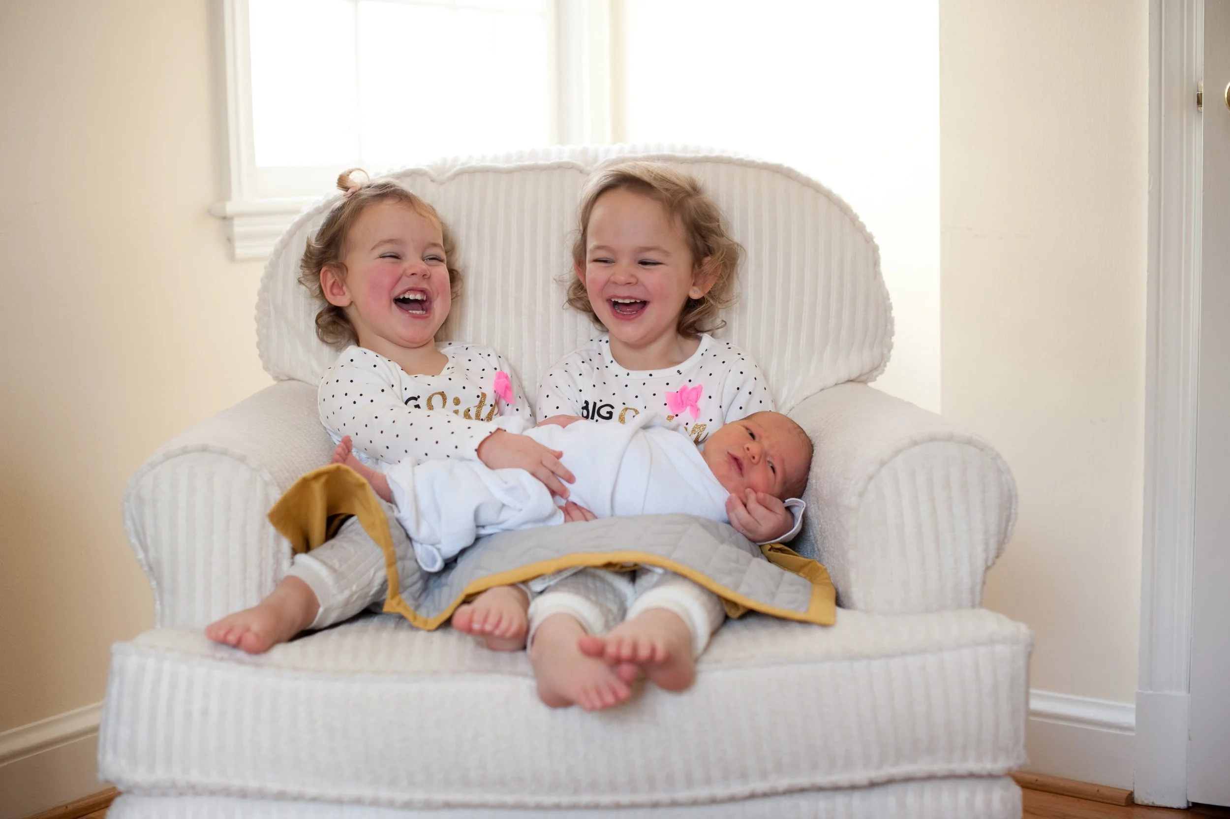 Two young girls sitting on a white armchair, smiling and laughing, while holding a newborn baby wrapped in a white blanket.