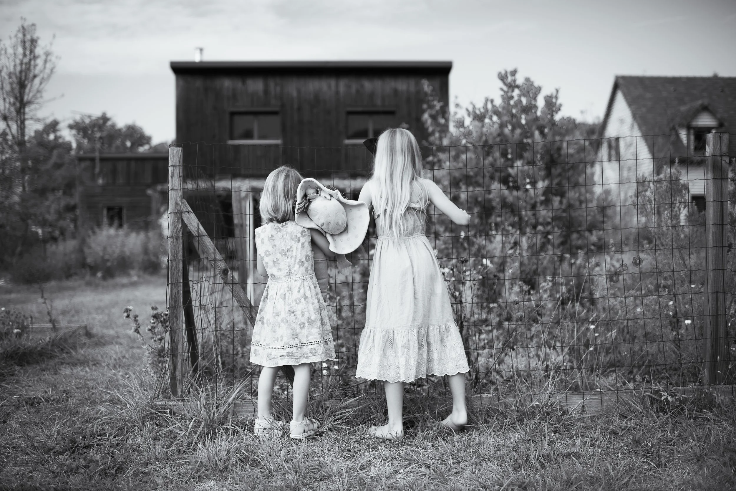 Two young girls with long hair, wearing dresses, stand in front of a wire fence and look toward a dark wooden house and neighboring house in a rural area, in a black-and-white photograph.