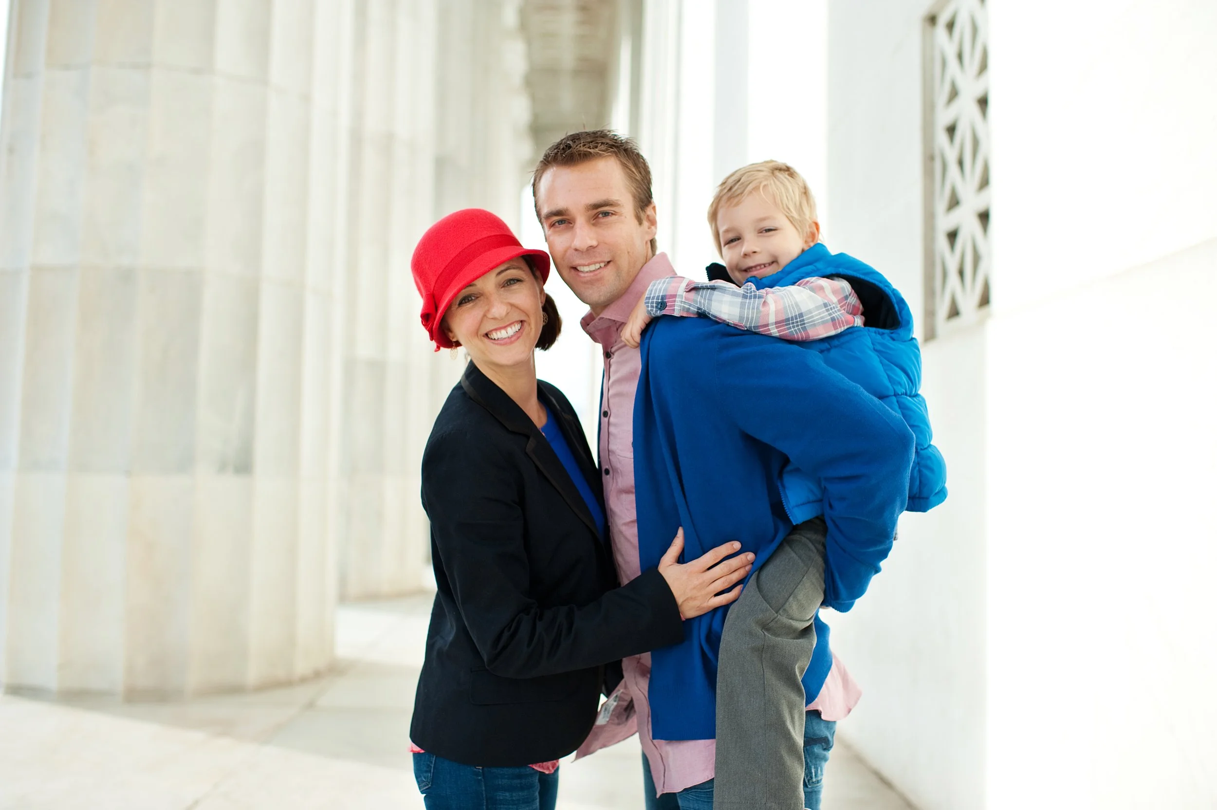 Family of three, smiling, standing outdoors on a sunny day. The woman wears a red hat and a black jacket, the man holds a young boy on his back. The boy is dressed in a blue jacket and is smiling. The background features a light-colored building with