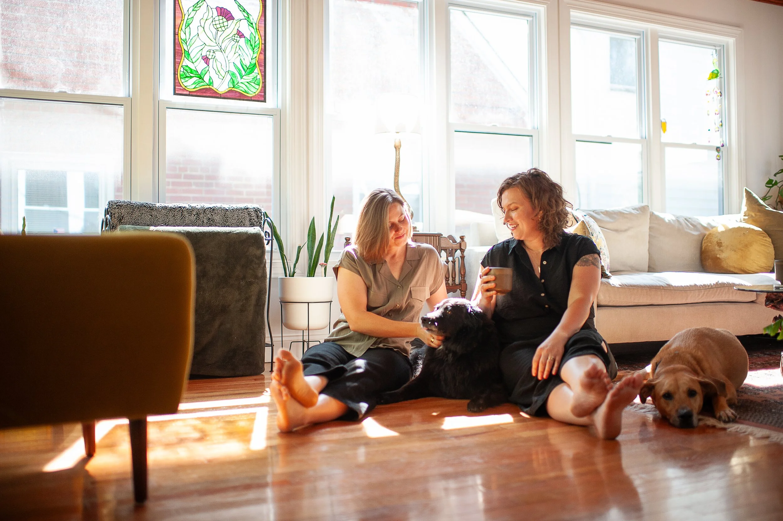 Two women sit on a wooden floor in a bright living room, smiling and petting a black dog. The room has large windows, a beige sofa with cushions, a potted plant, and colorful decorations. One woman has short blonde hair, and the other has curly brown