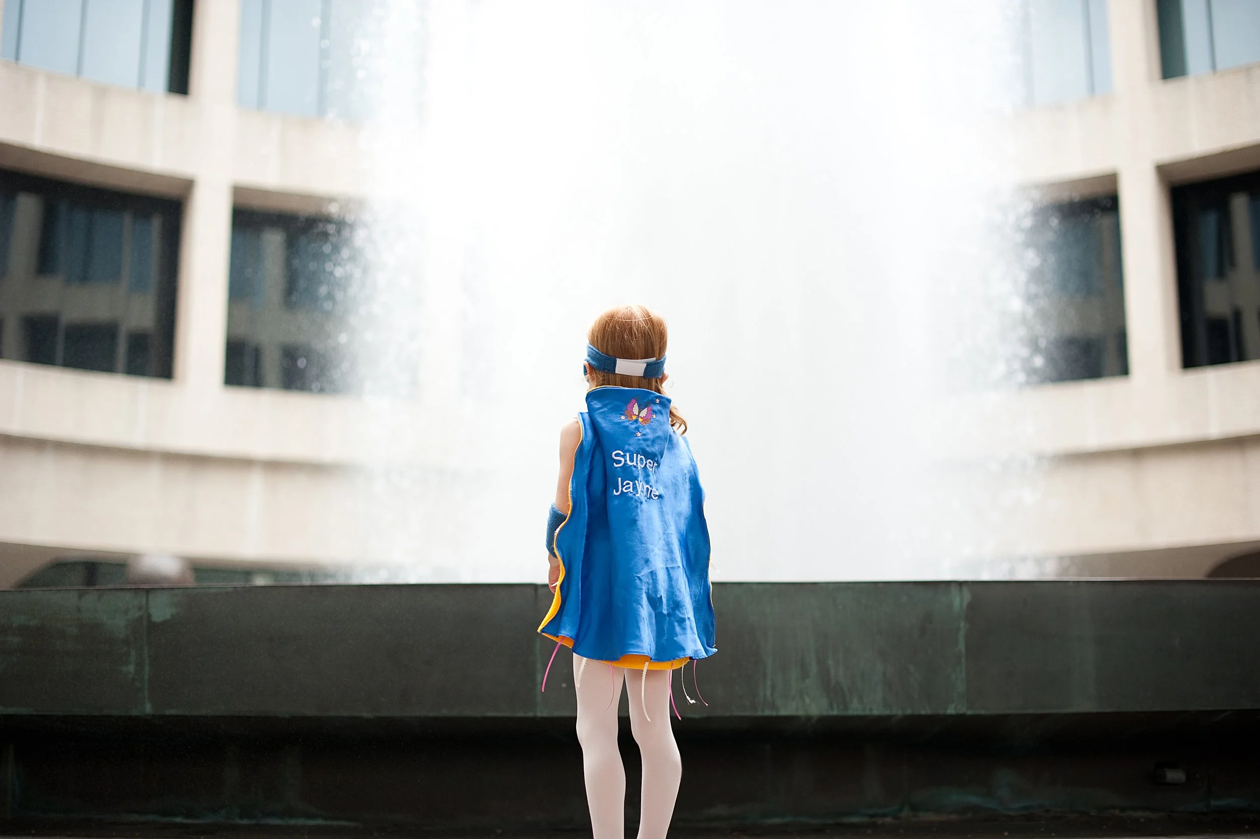A young girl in a superhero costume with a cape, standing in front of a large fountain in a modern building atrium, with her back to the camera.