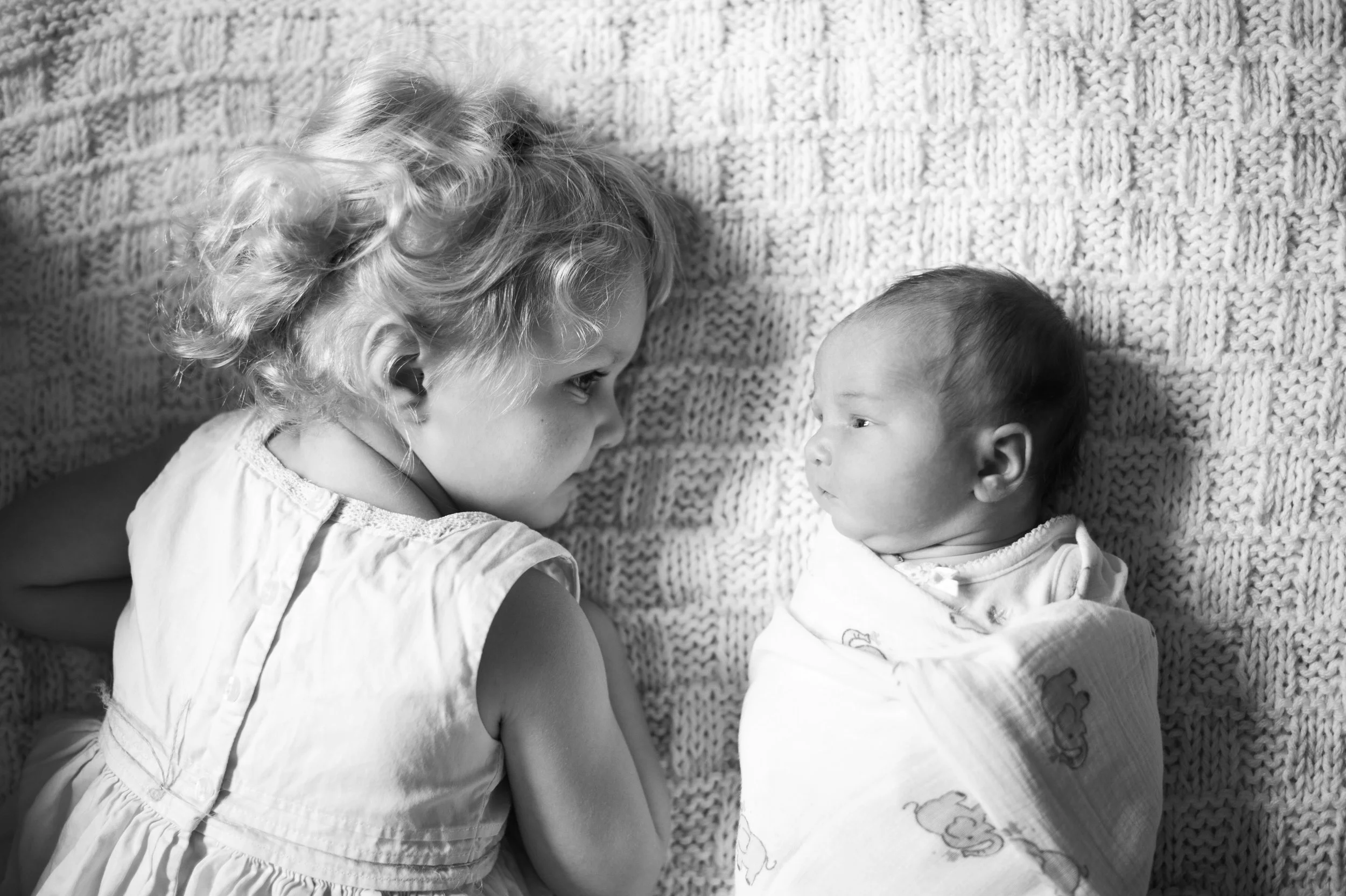 A young girl and an infant lying on a textured blanket, facing each other.