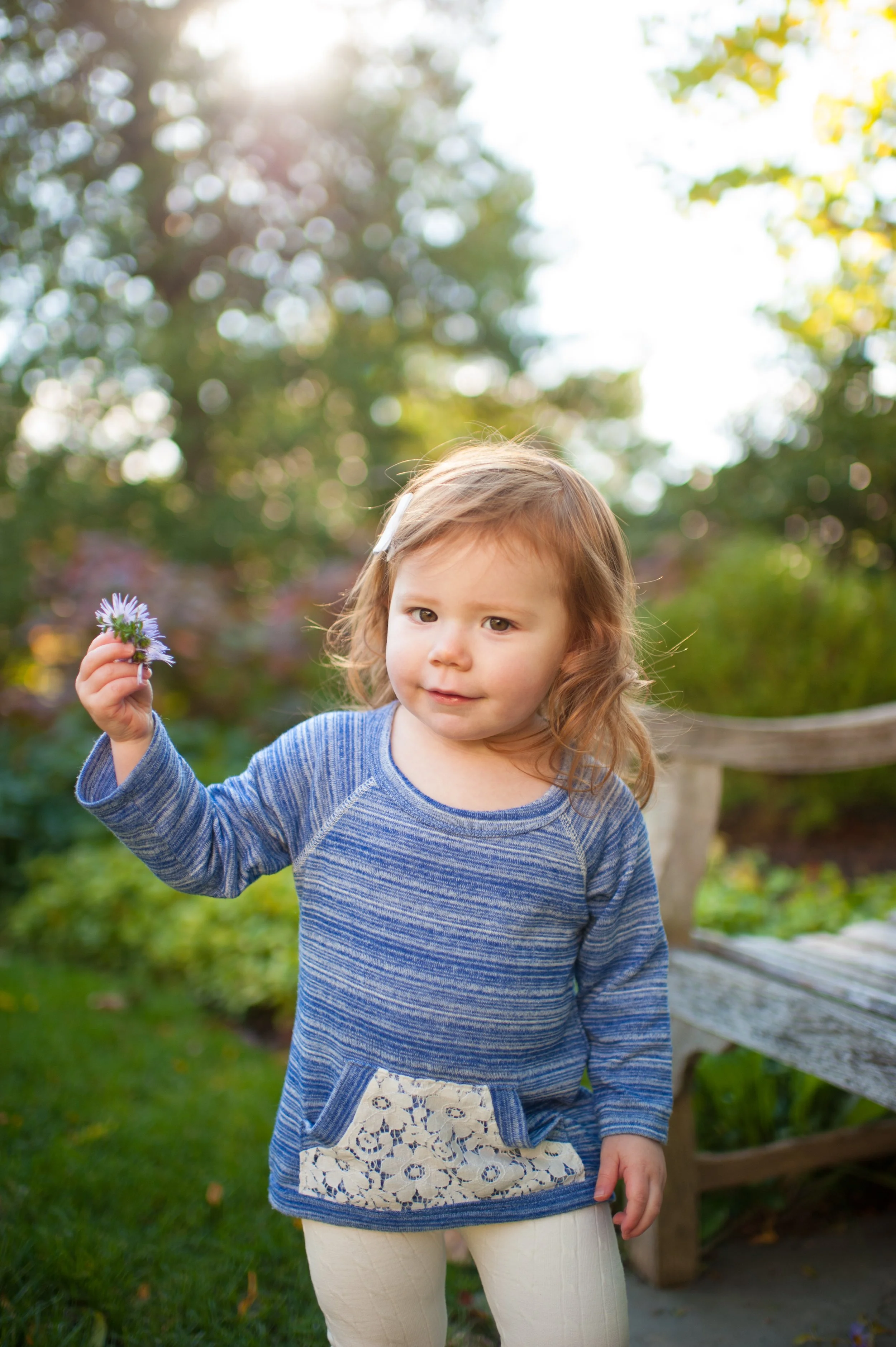 A young girl with light brown hair and a white hair clip holds a small purple flower in her hand outdoors at sunset, with trees and a wooden bench in the background.
