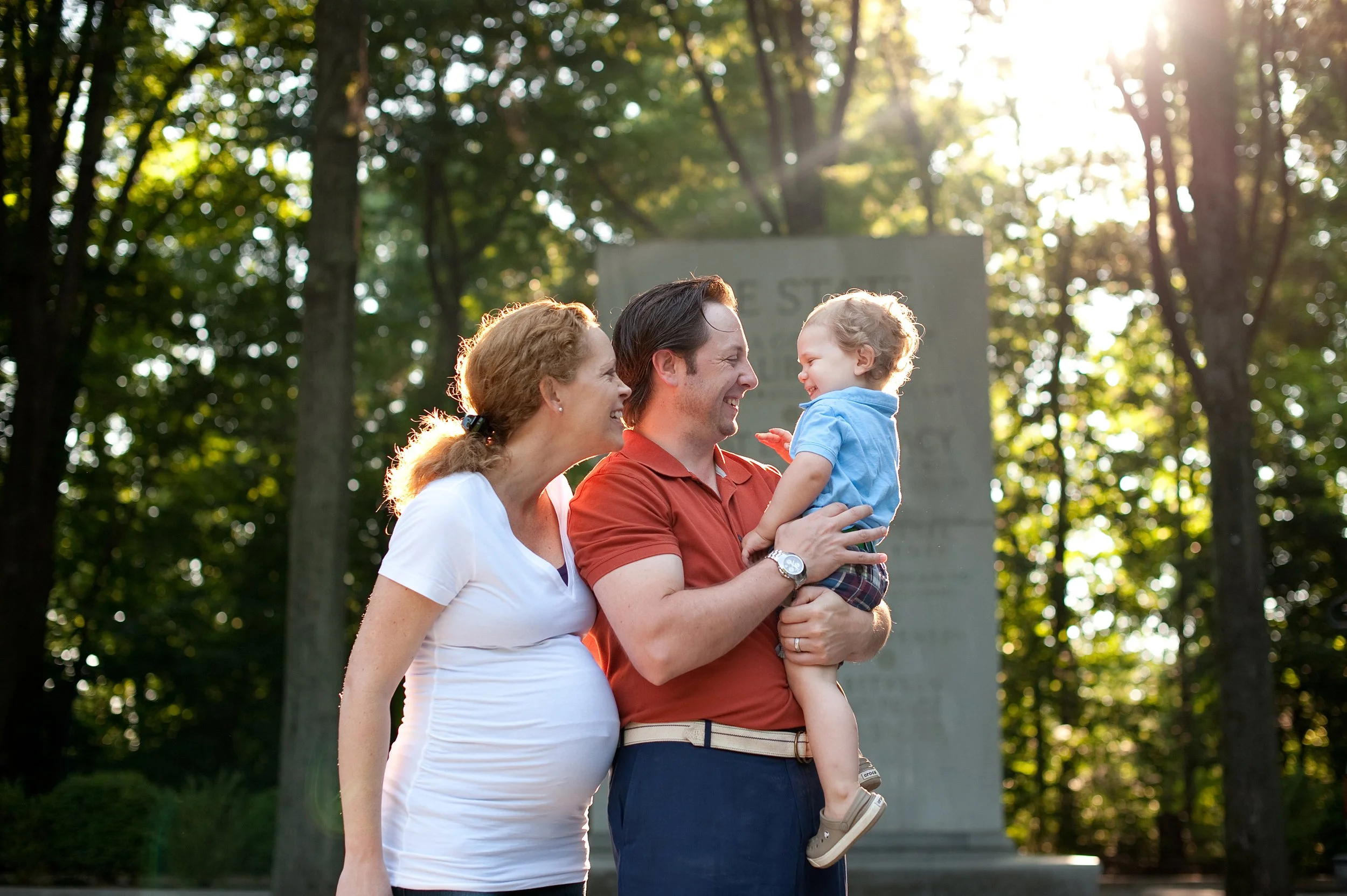 A family of three, including a pregnant woman, a man, and a young boy, are smiling and looking at each other outdoors in a park during sunlight, with trees in the background.