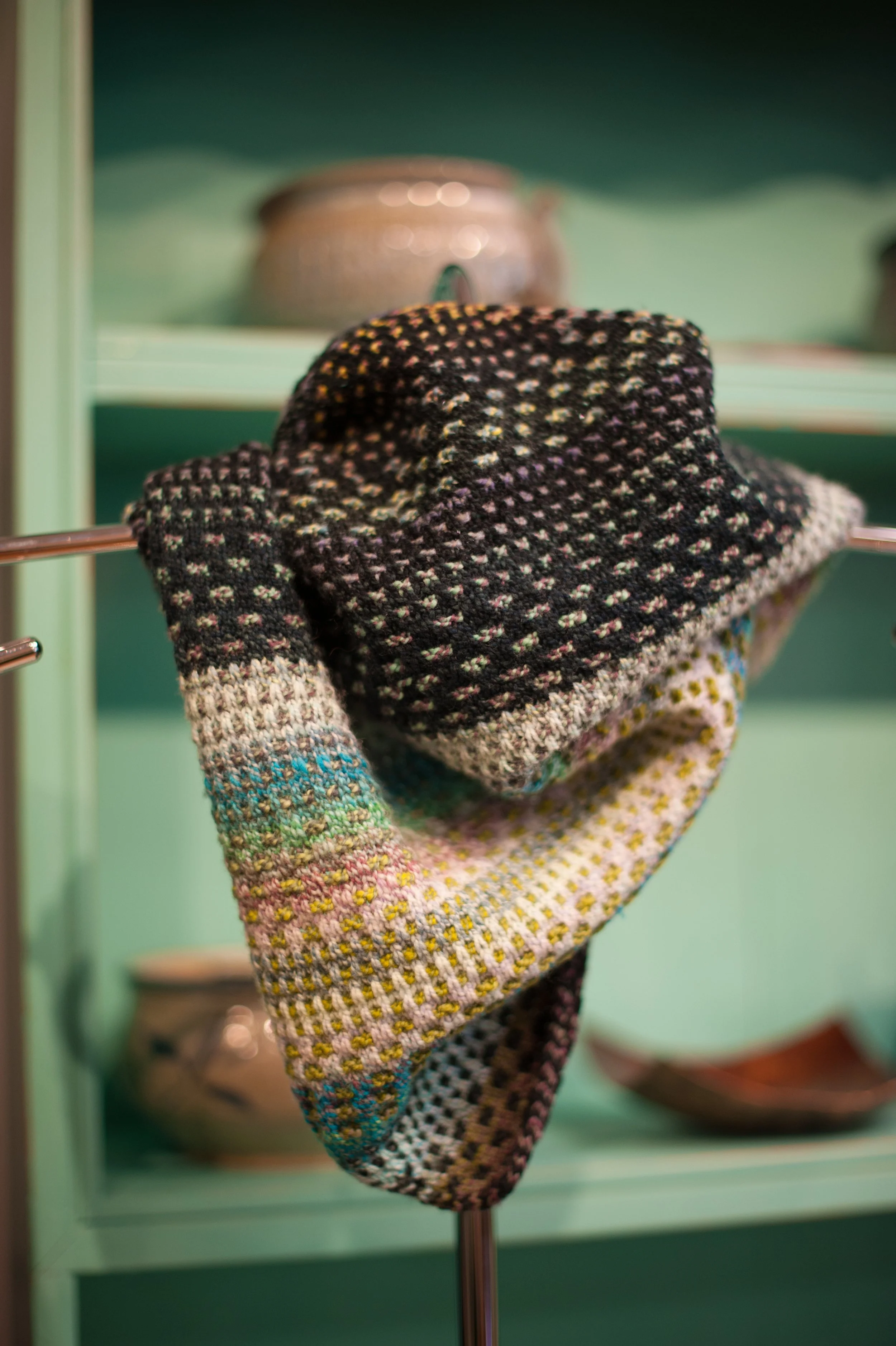 Colorful woven scarf with black, white, yellow, pink, and blue patterns hanging on a metal rod in front of a painted teal background, with ceramic pots on shelves behind.