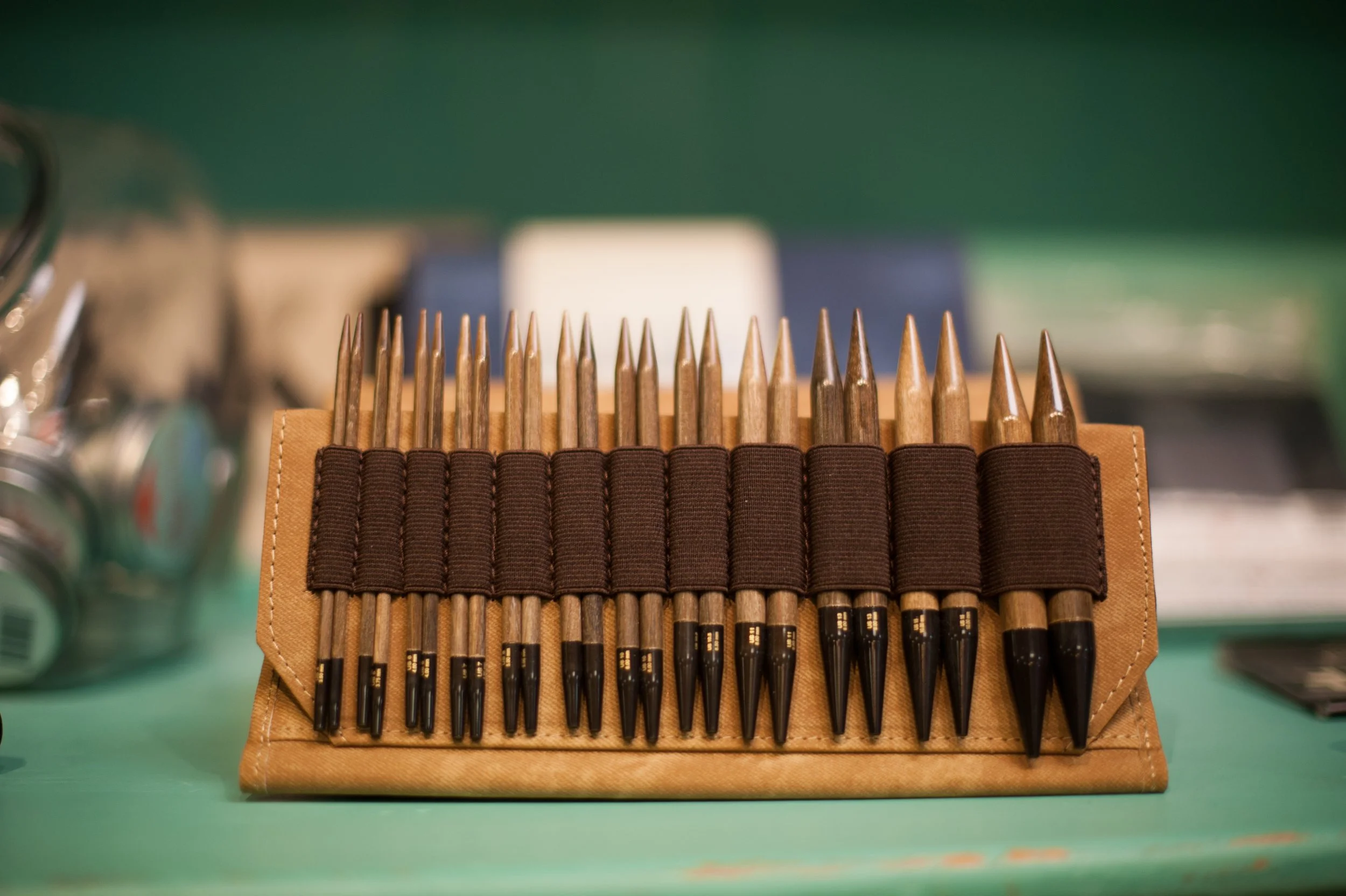 A row of 15 knitting needles with black tips, held in a tan fabric case with elastic loops, on a green surface.