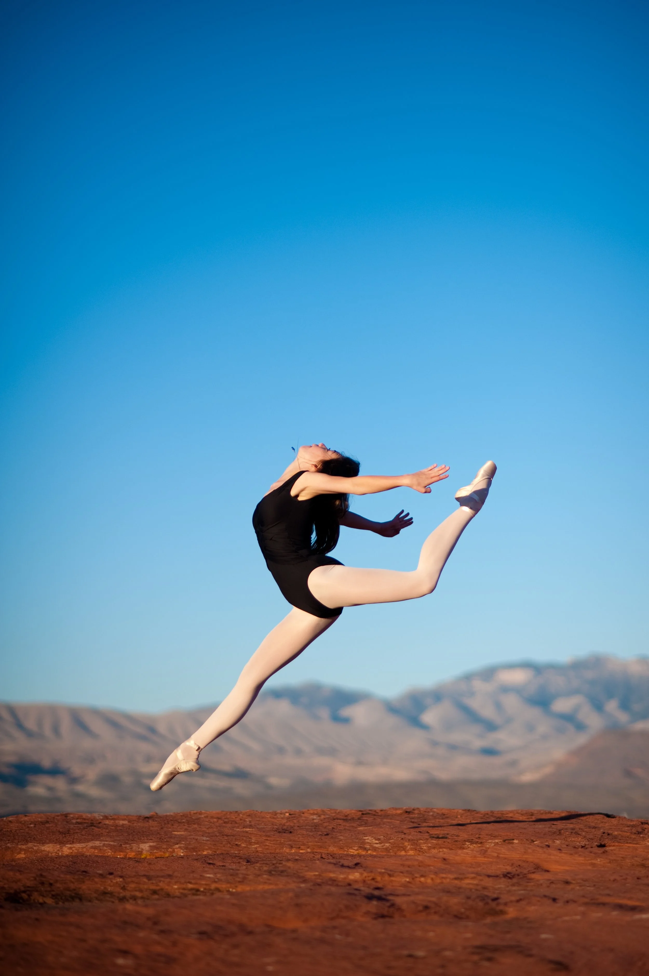 Ballet dancer in mid-air leap on red rocky ground with mountains and clear blue sky in the background.