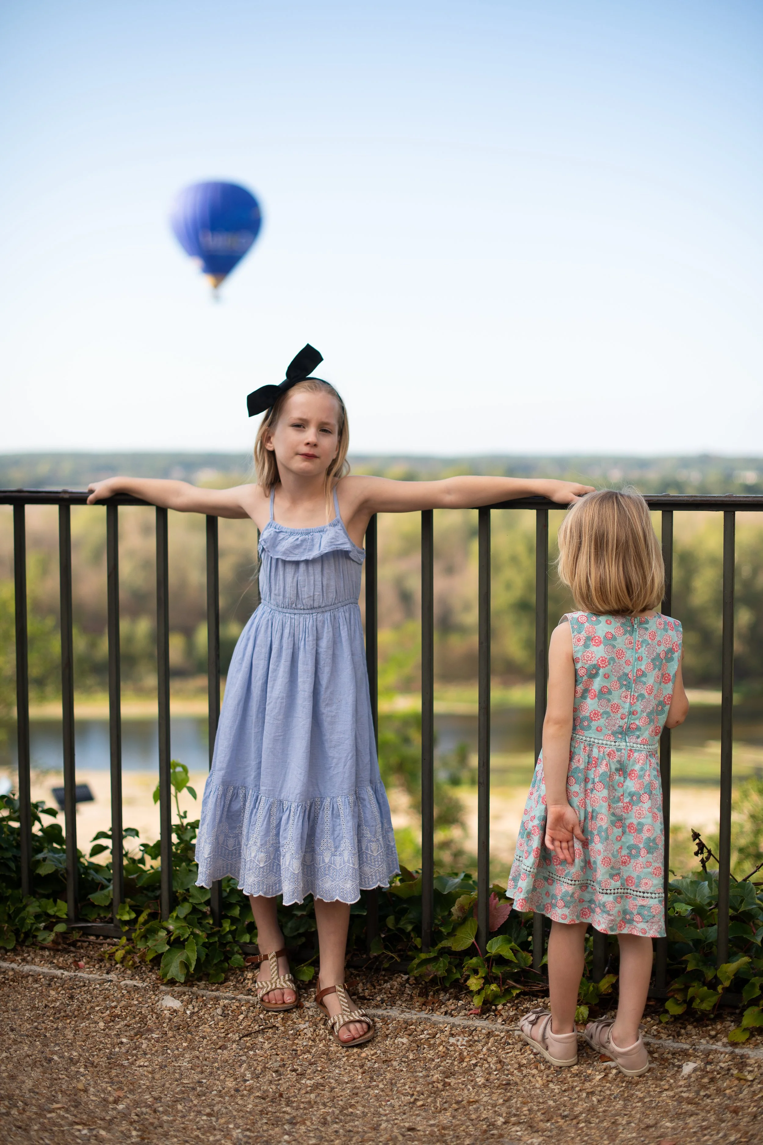 Two young girls standing by a black metal railing on a hill, with a blurry hot air balloon in the sky in the background. One girl is wearing a blue dress with a black bow in her hair, and the other girl has a floral dress and is facing away.