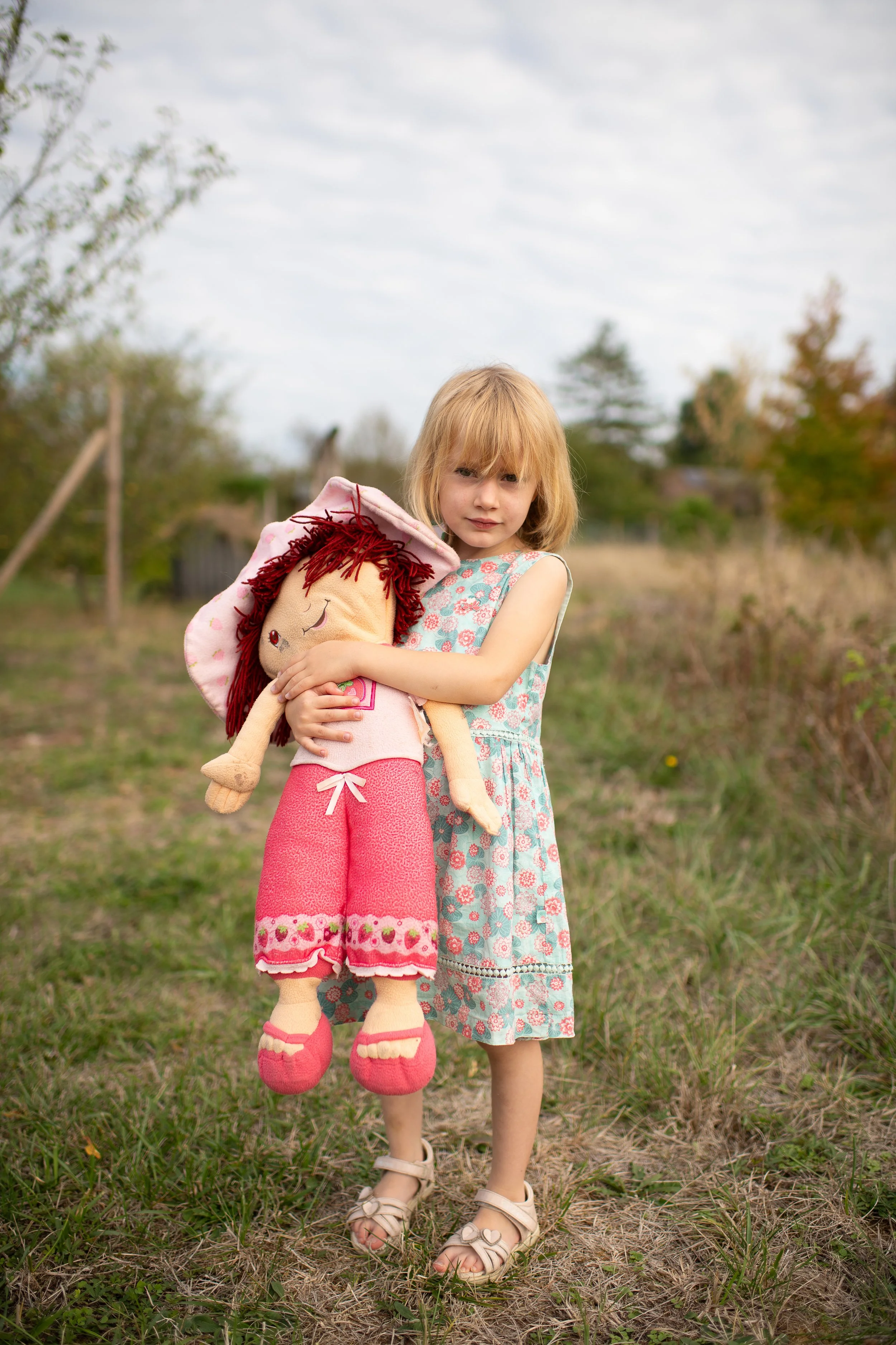 A young girl with blonde hair stands outdoors on a grassy area, holding a large plush doll with red hair and a pink hat. She is wearing a light blue dress with a floral pattern and beige sandals. The background features a slightly overcast sky, trees