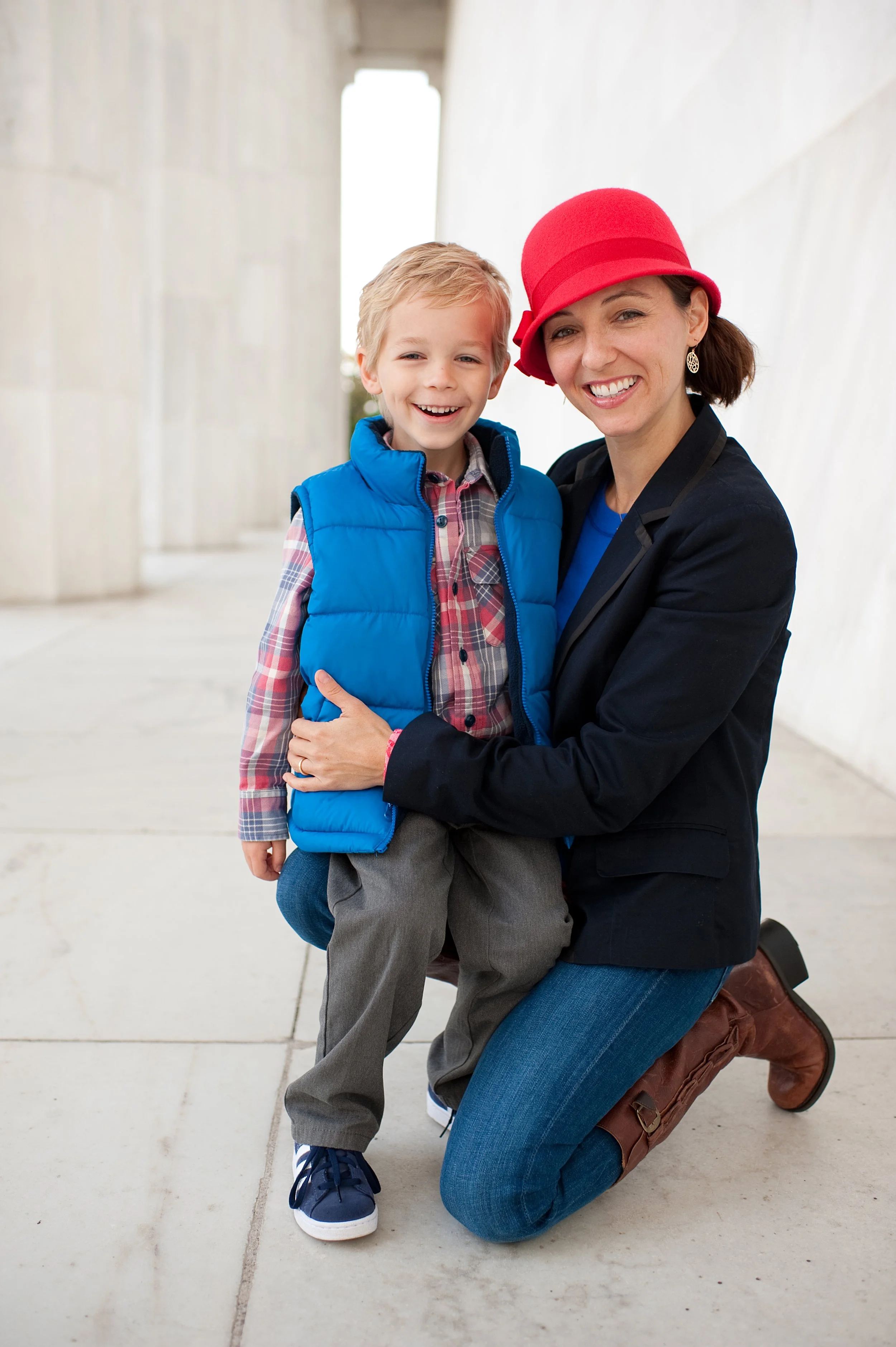 A woman kneeling outdoors, holding a young boy, both smiling, dressed casually, with an architectural structure in the background.