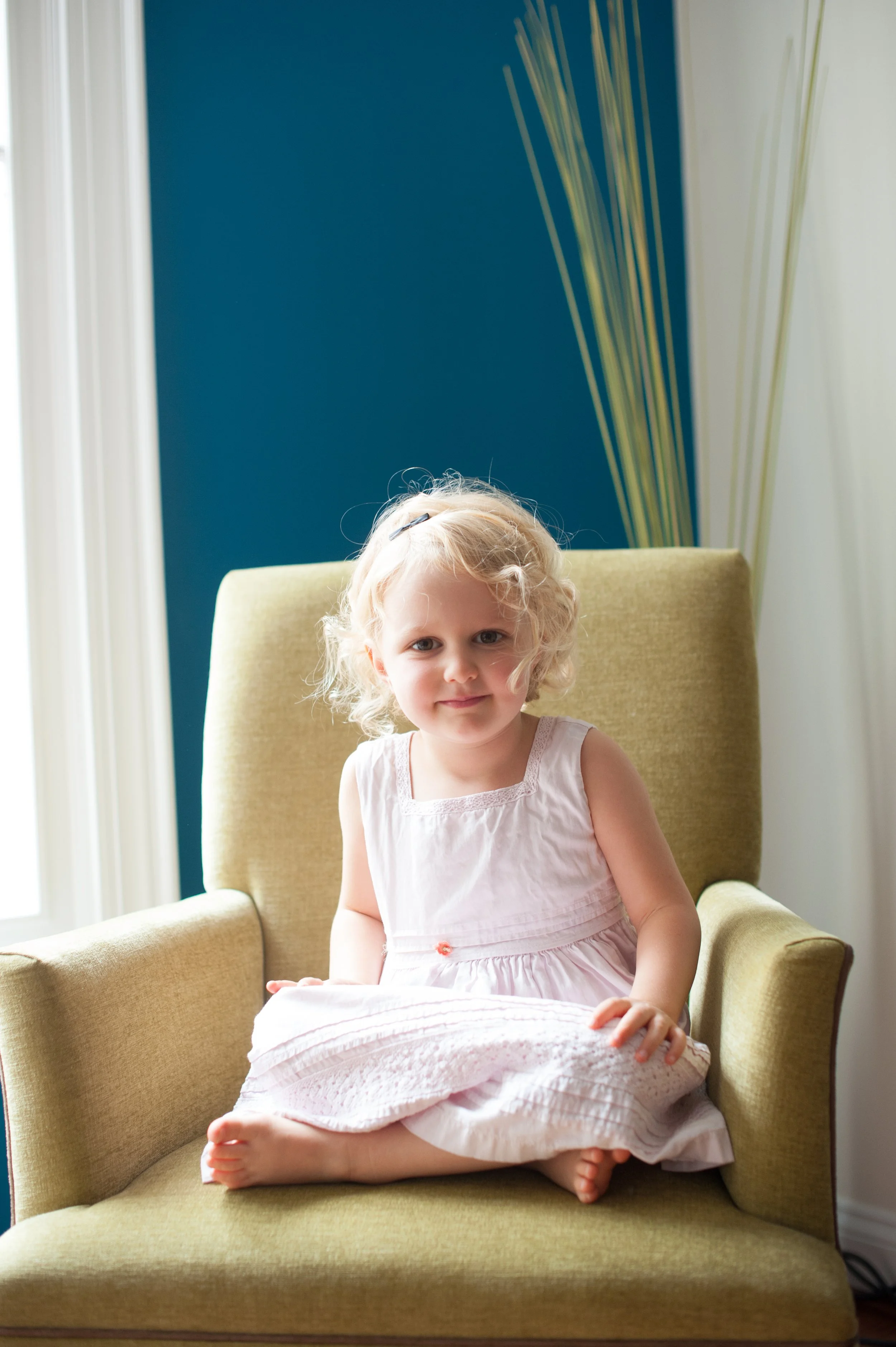 A young girl with curly blonde hair sitting cross-legged on an olive green armchair, wearing a light pink sleeveless dress, indoors near a window with white curtains, and a blue accent wall with decorative reeds in the background.
