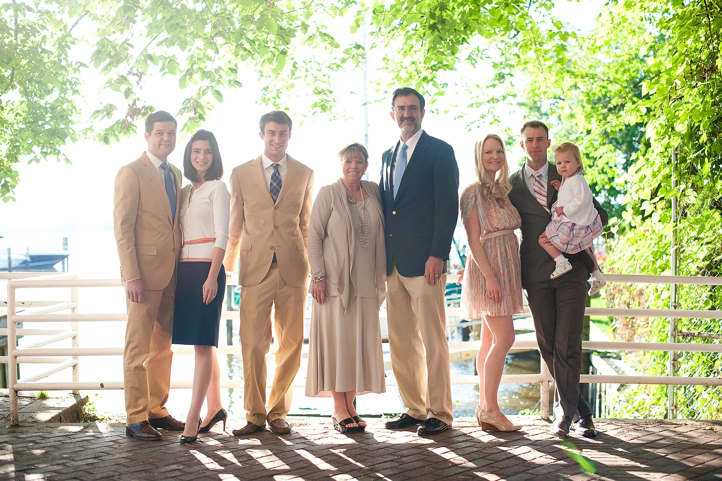 A family of eight standing outdoors near water, smiling, with green trees overhead.