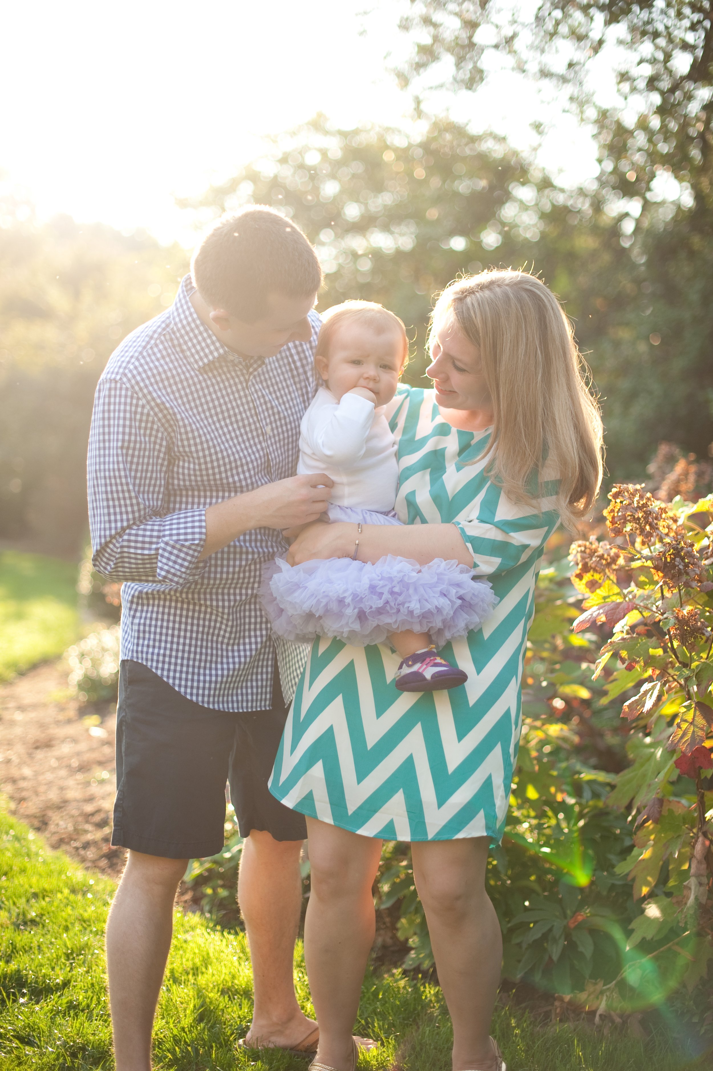 A family of three, a father, mother, and their young daughter, standing outside in sunlight among greenery and bushes, smiling and enjoying time together.