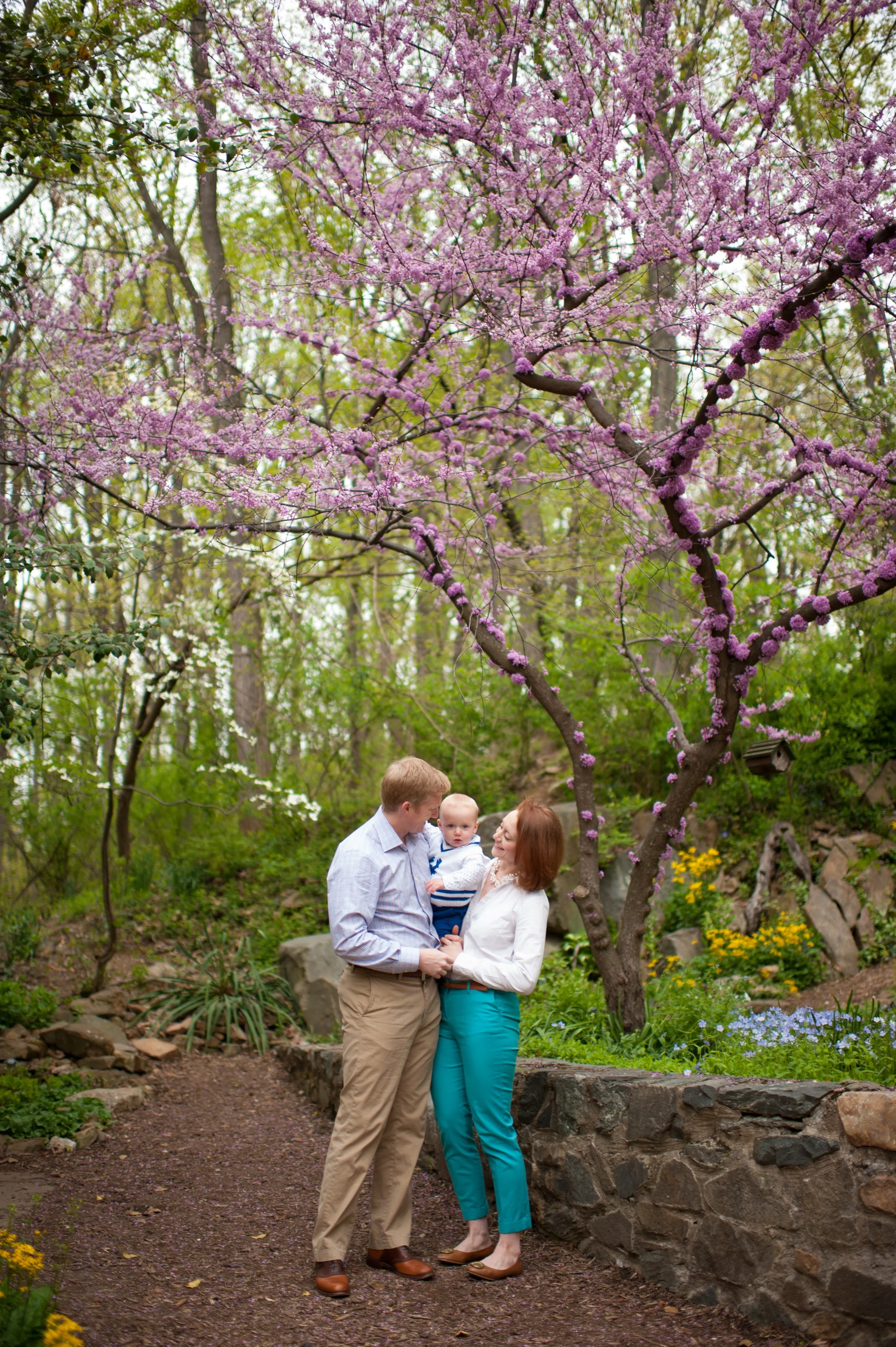 A family of three, a man, woman, and a baby, standing in a garden with blooming trees and colorful flowers.