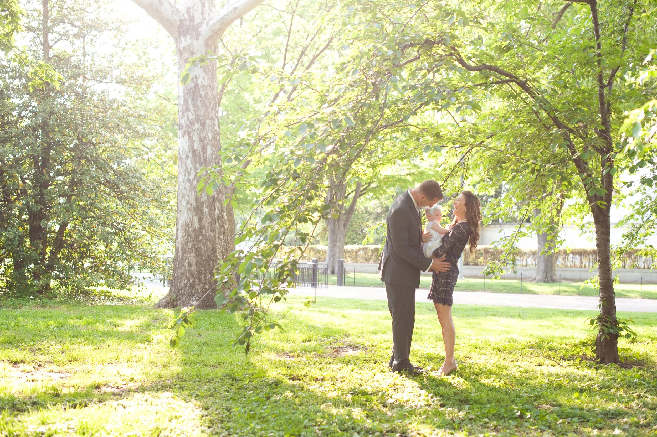 A family of three enjoying a moment in a park with lush green trees and grass, sunlight filtering through the leaves, with a man, woman, and a baby.