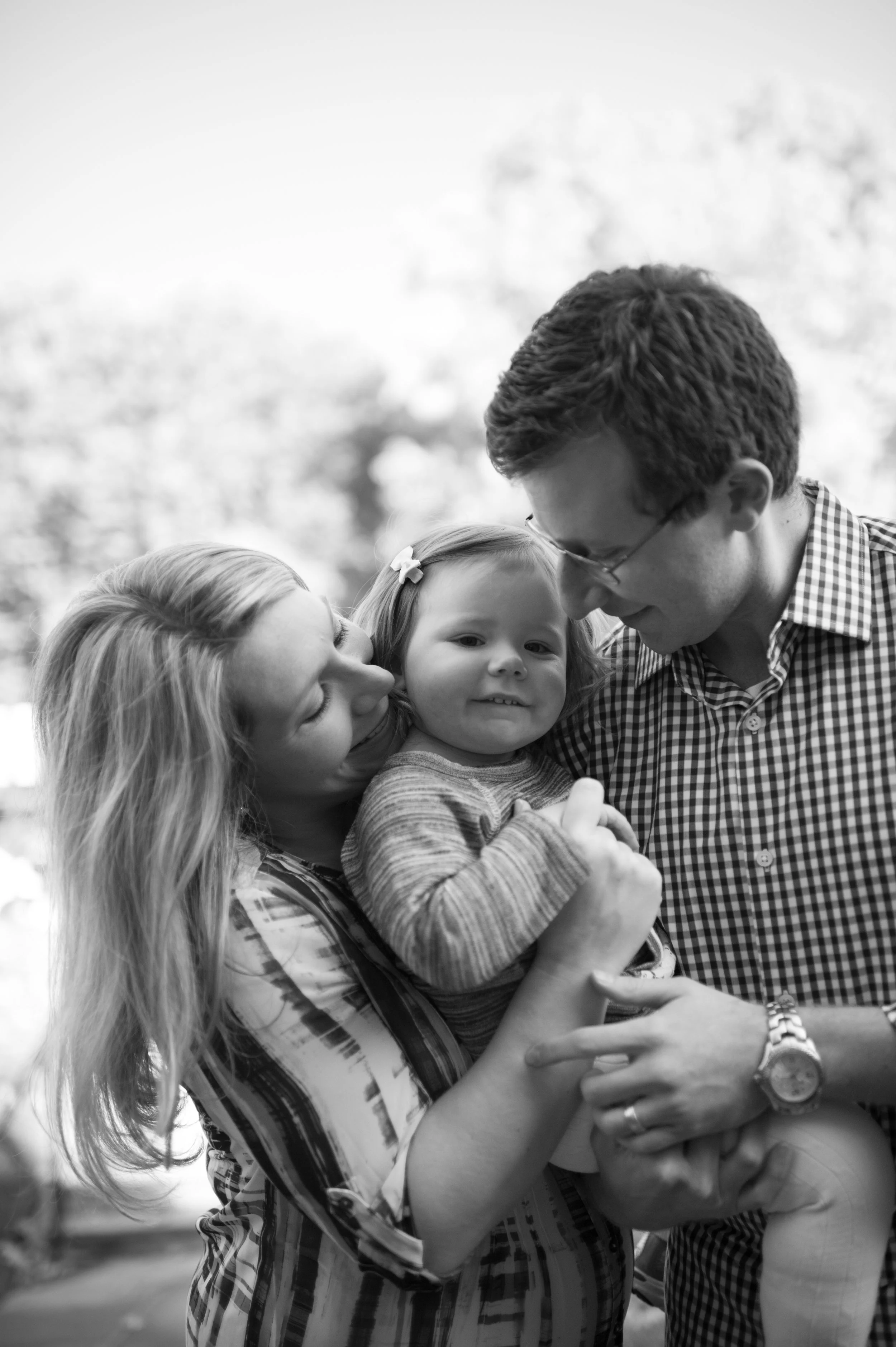 A smiling woman holding a young girl, with a man leaning in close, outdoors in a park. The scene is in black and white.
