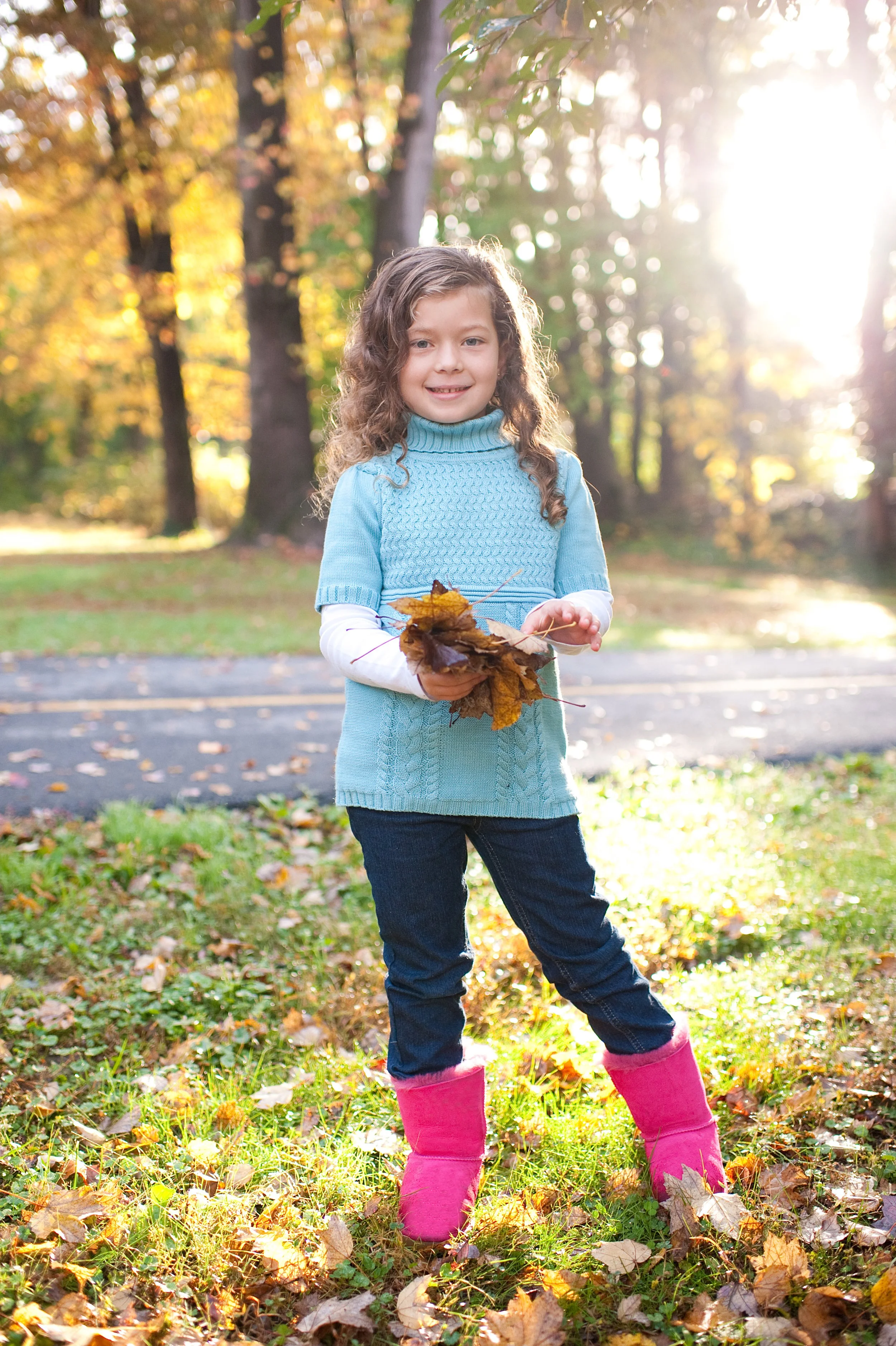 A young girl standing on a grassy patch of ground covered with fallen autumn leaves, holding a bunch of dried leaves, wearing a light blue sweater, dark jeans, and bright pink boots, with trees and sunlight in the background.
