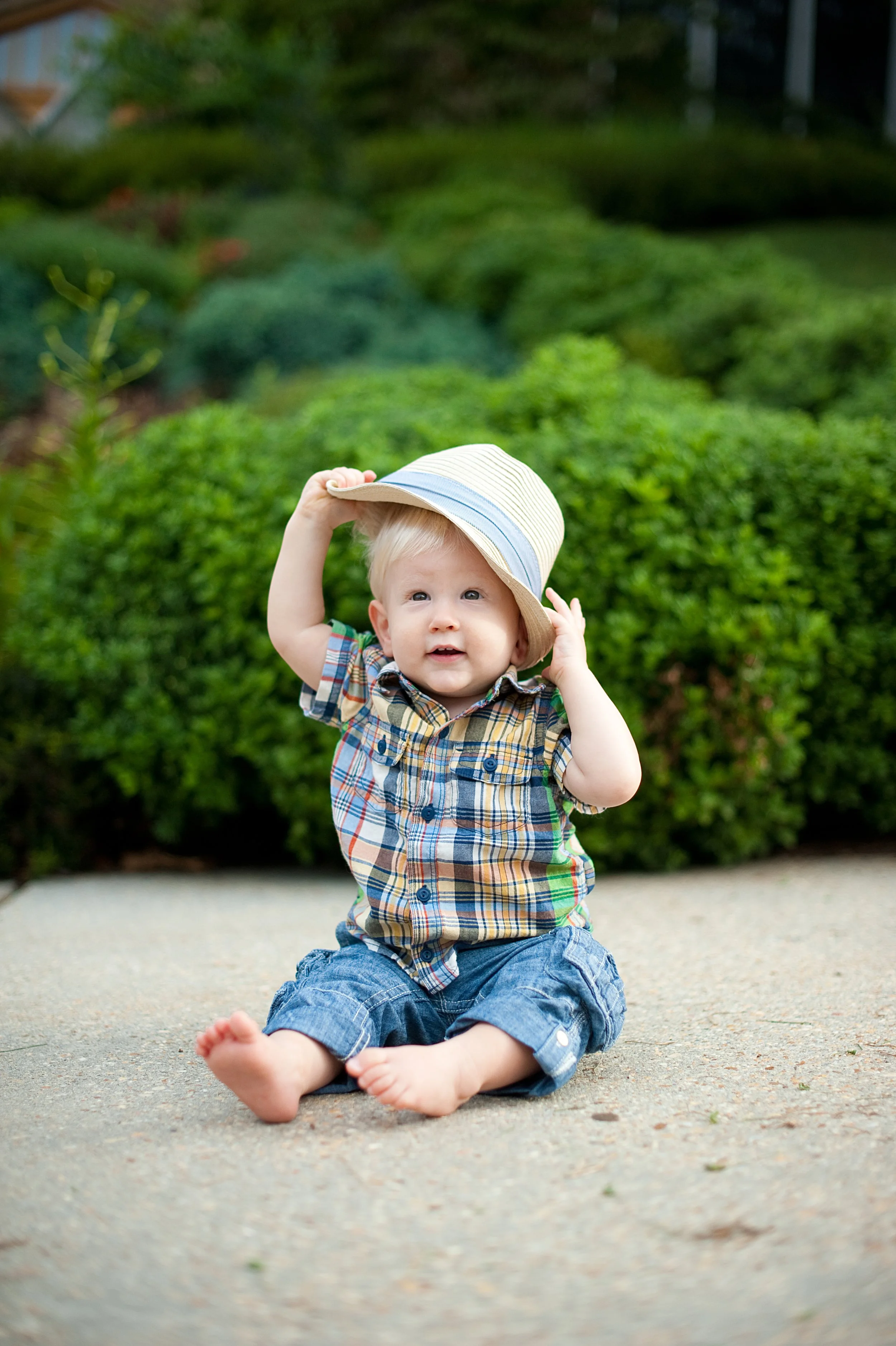 A young child sitting on a sidewalk, holding a straw hat, dressed in a plaid shirt and denim shorts, with lush green bushes in the background.