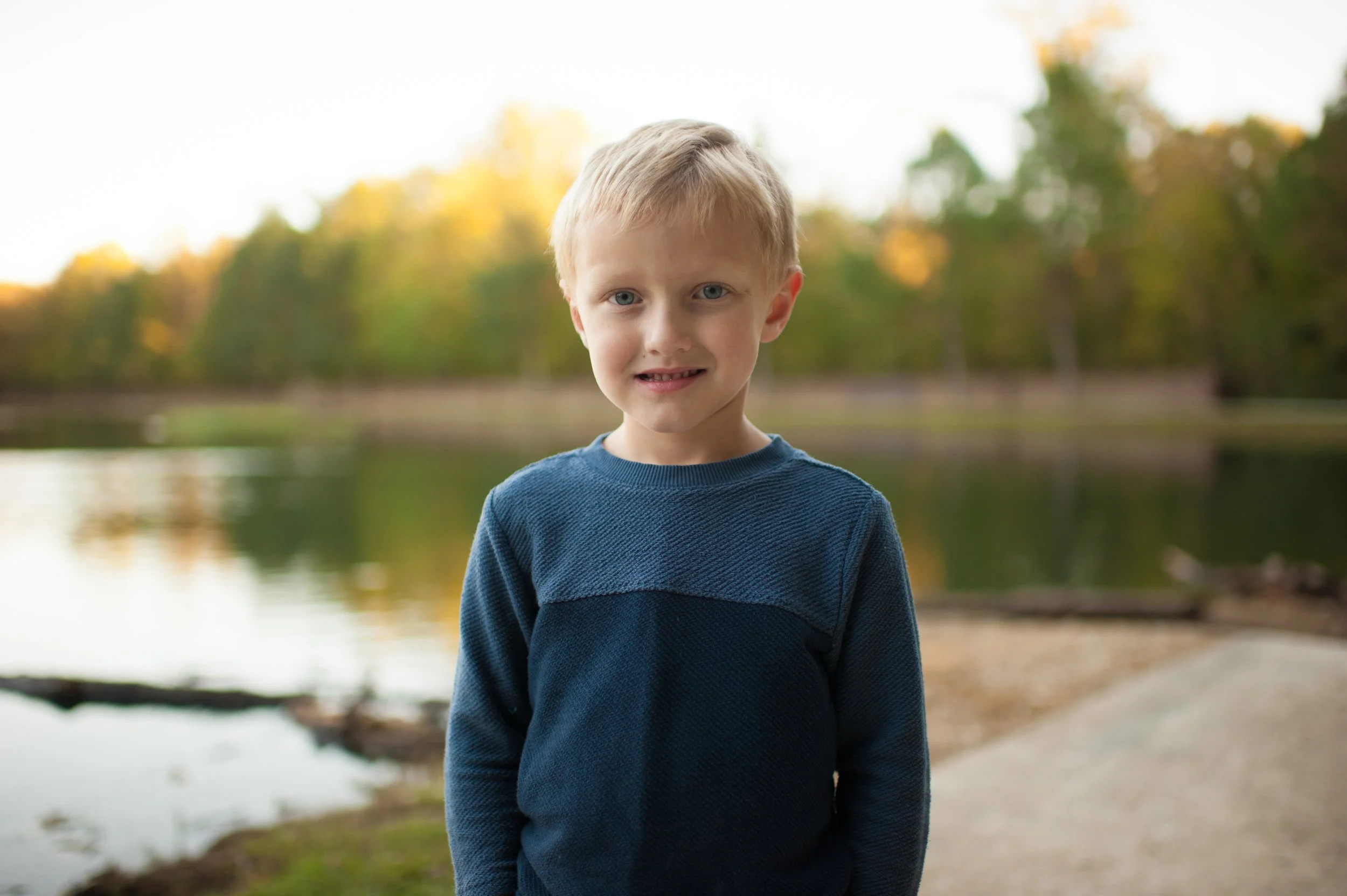 A young boy with blonde hair and blue eyes standing near a lake in a park during fall with trees showing autumn colors.