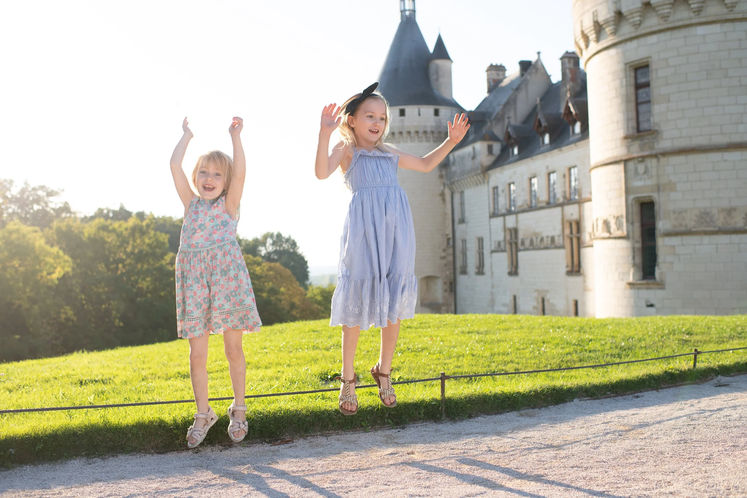 Two young girls in dresses jumping on a small fence in front of a castle, with trees and a clear sky in the background.
